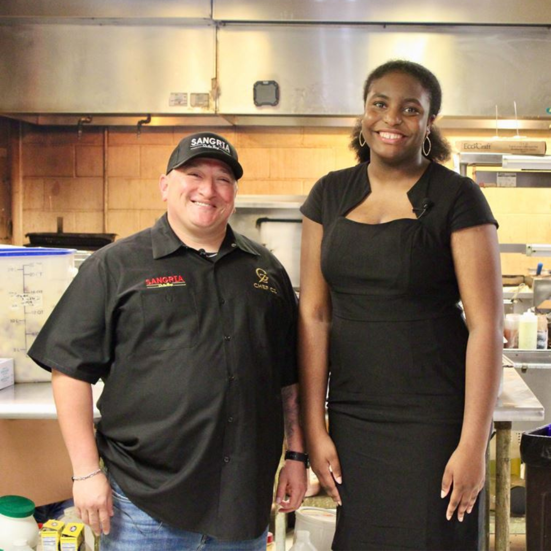 A chef in a black uniform and cap stands next to a person in a black dress in a commercial kitchen.