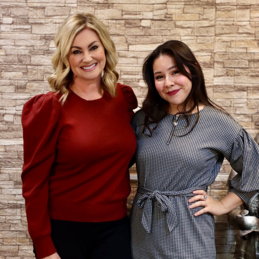 Two people standing side-by-side smiling in front of a textured stone-patterned wall.