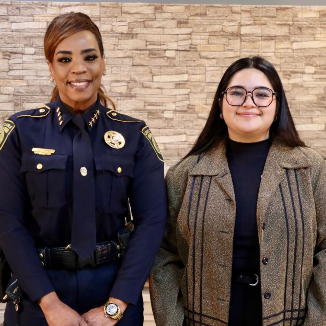 A police officer in uniform stands next to a person in a brown blazer, both smiling against a light stone-textured wall.