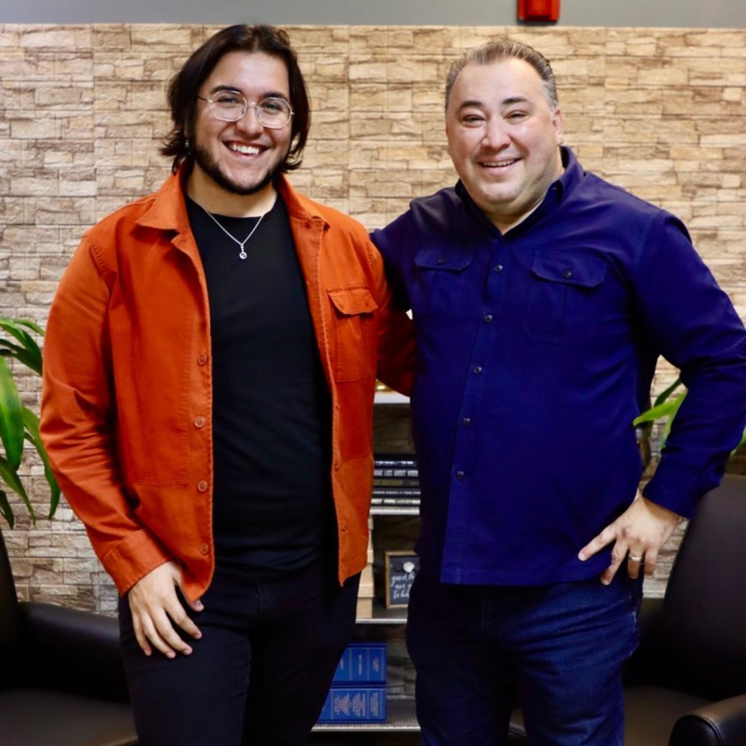 Two smiling people stand side-by-side, one wearing an orange jacket and the other a blue button-down shirt, near a bookshelf.
