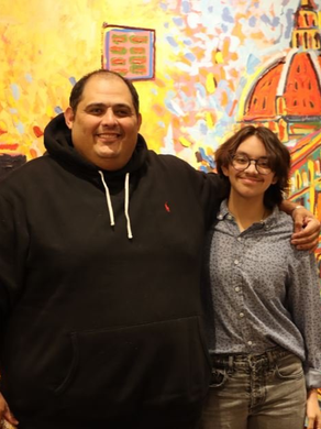 A smiling person in a black hoodie and a smiling person in a patterned shirt stand together before an abstract mural.