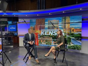 A man in a suit and a woman in a dress sit in director's chairs on a KENS 5 television news studio set.