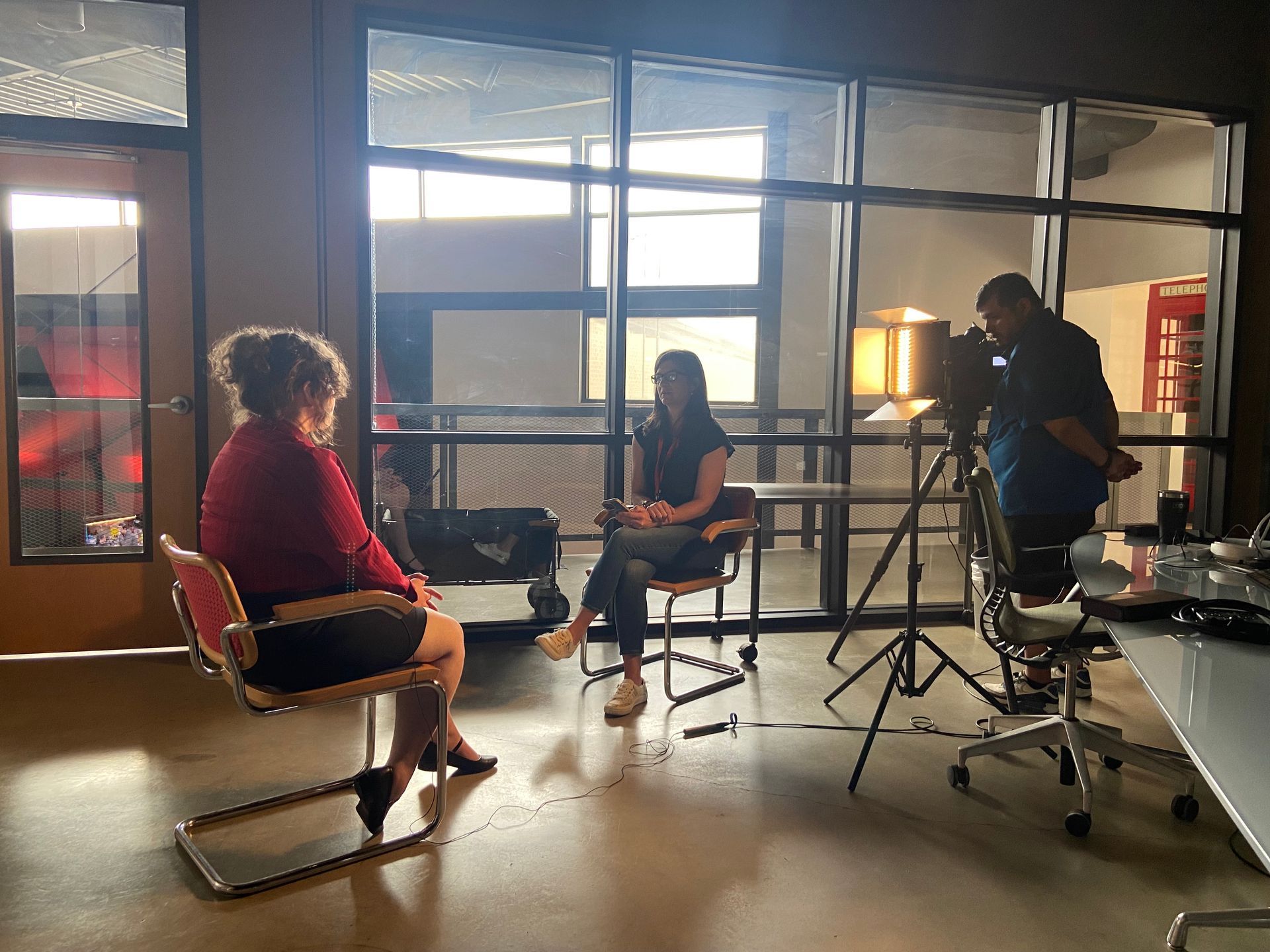 A person being interviewed by another individual while a camera operator films them in a sunlit office space.
