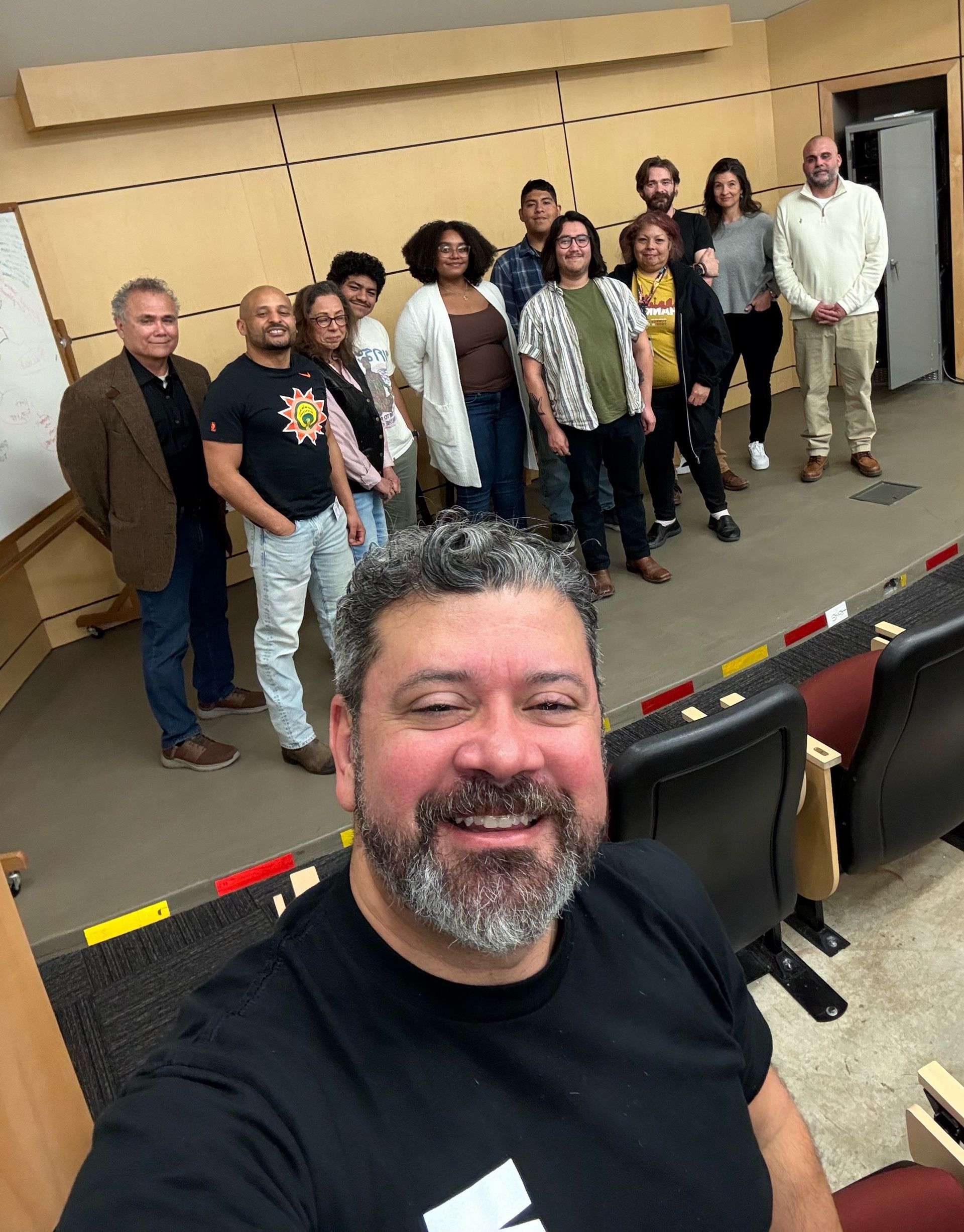 A man smiles while taking a selfie in a classroom with a group of people standing in the background.