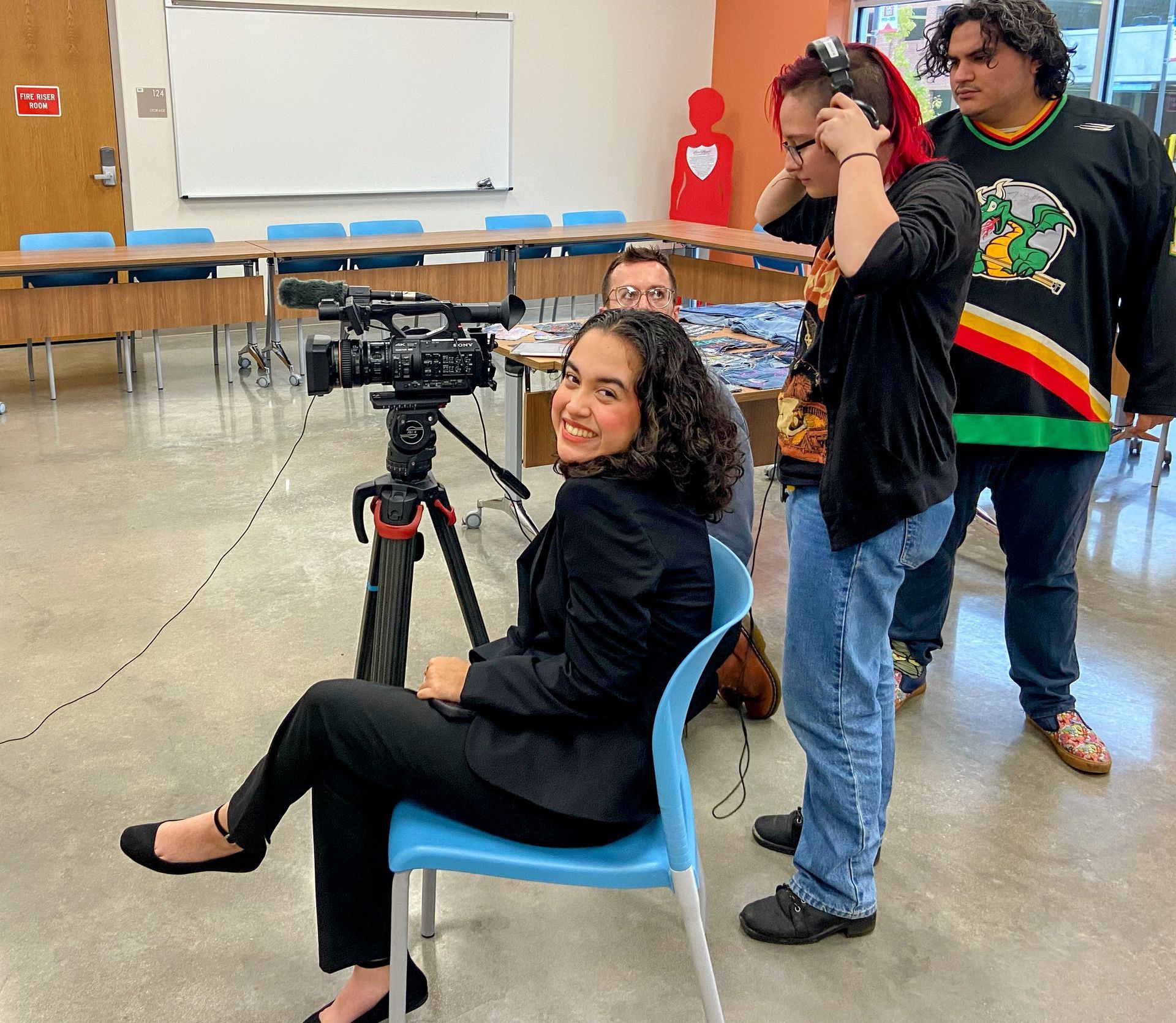 A person in a black suit sits at a video camera on a tripod, smiling, with two people standing behind them in a classroom.
