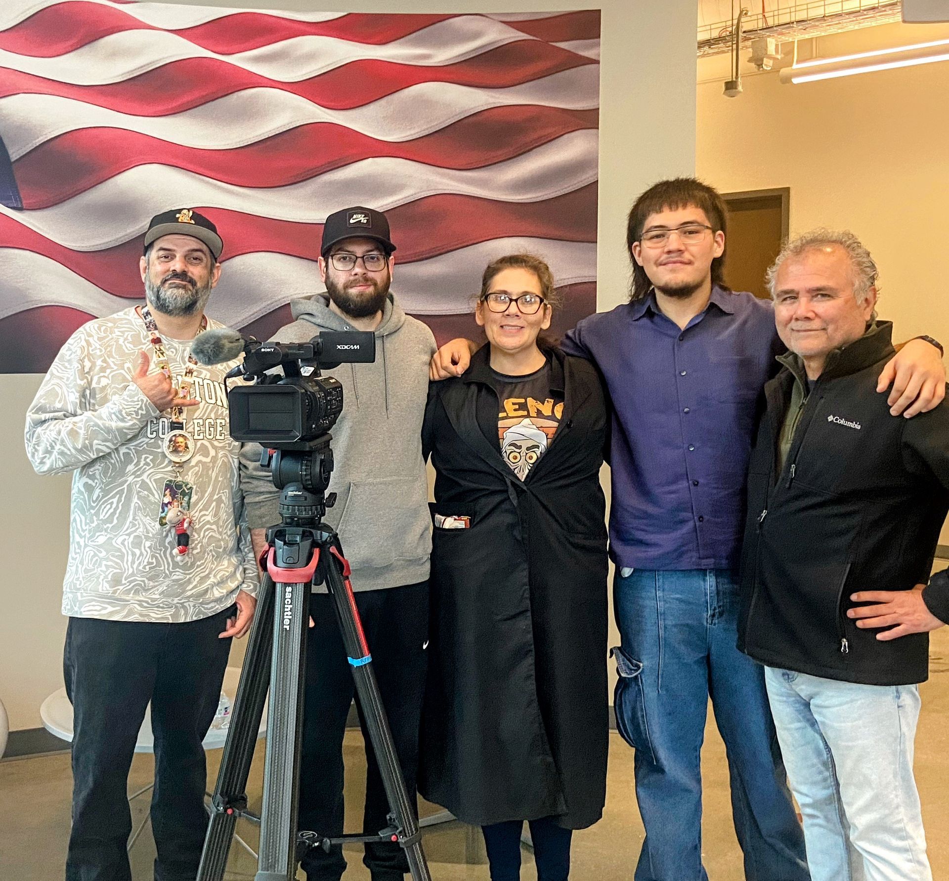 Five people stand in front of an American flag wall mural, with a video camera on a tripod positioned in the foreground.