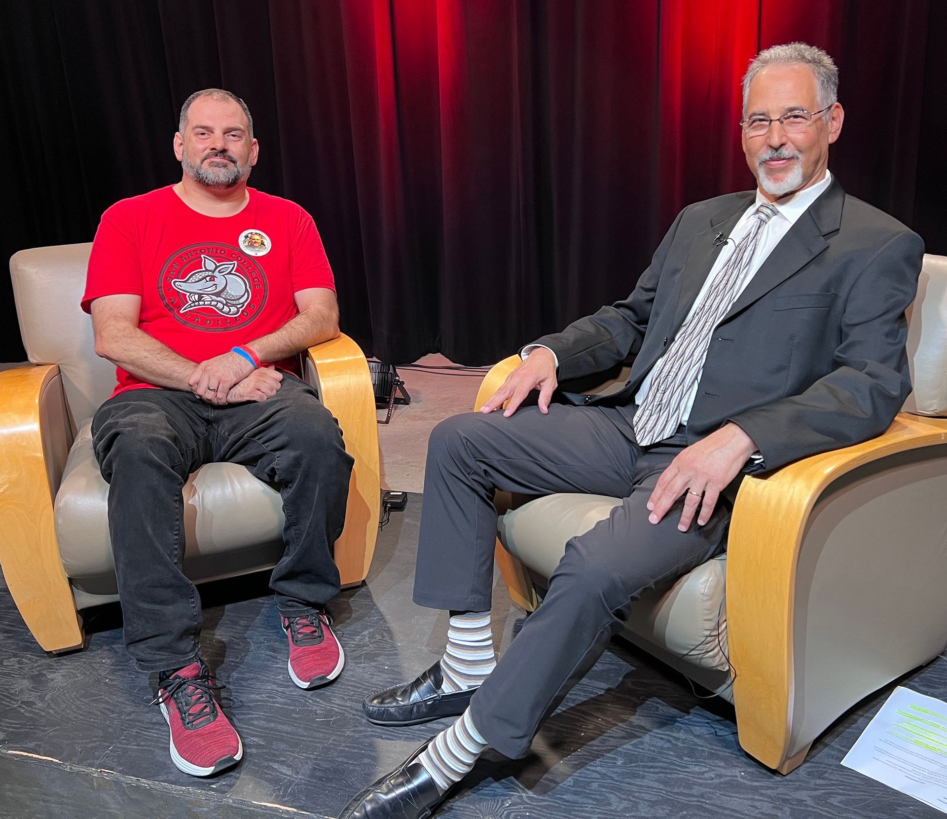 Two men sit in chairs on a stage with a red curtain backdrop; one wears a red t-shirt, the other a suit and tie.