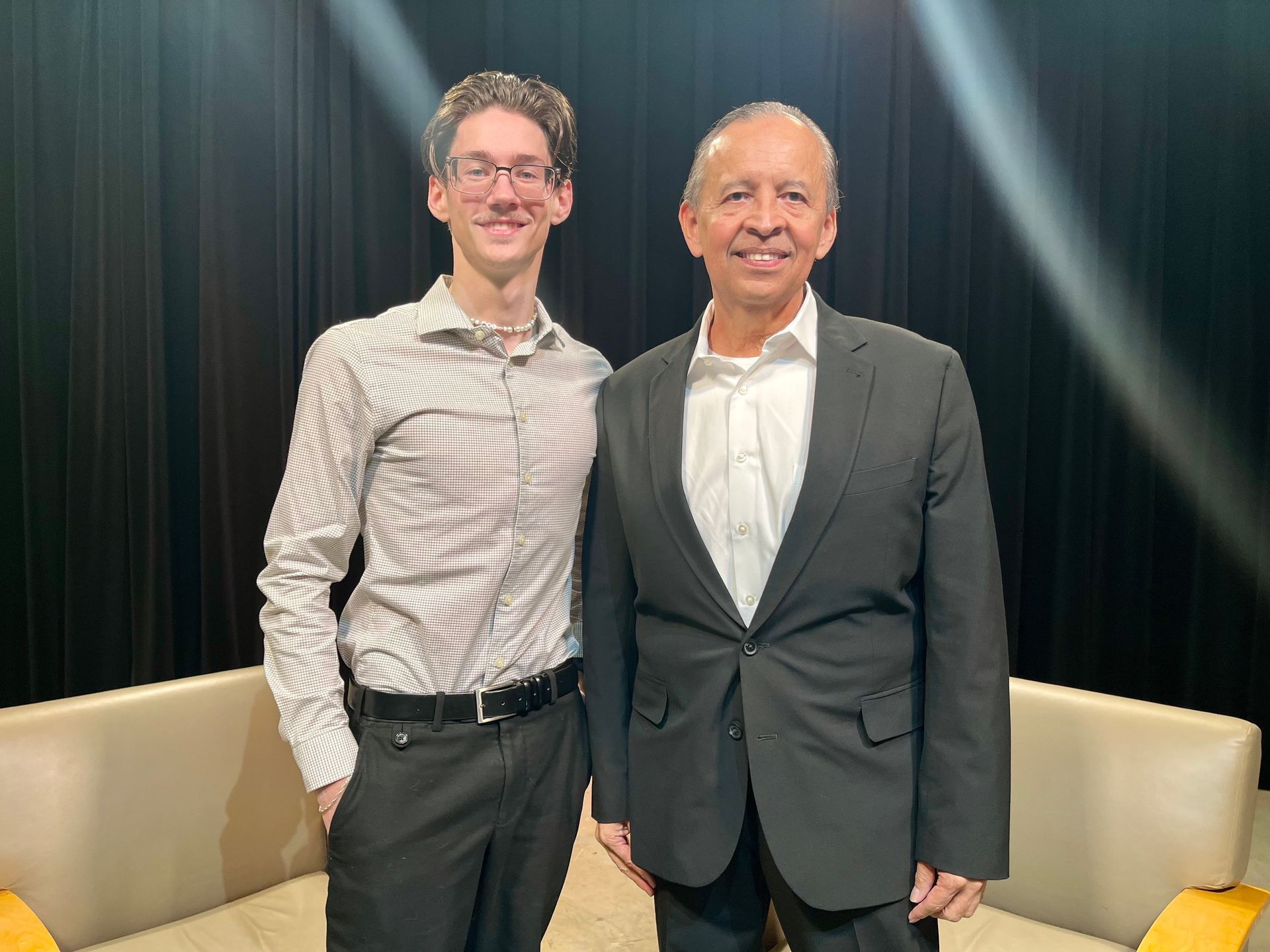 Two men standing together in front of a dark curtain backdrop, both smiling and wearing business casual attire.