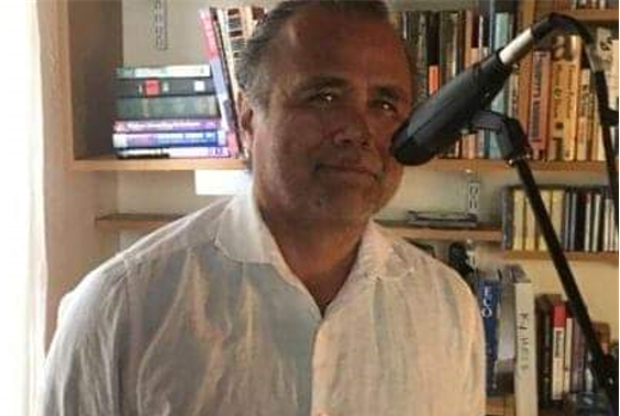 A person in a white button-down shirt stands in front of bookshelves with a microphone positioned near their face.