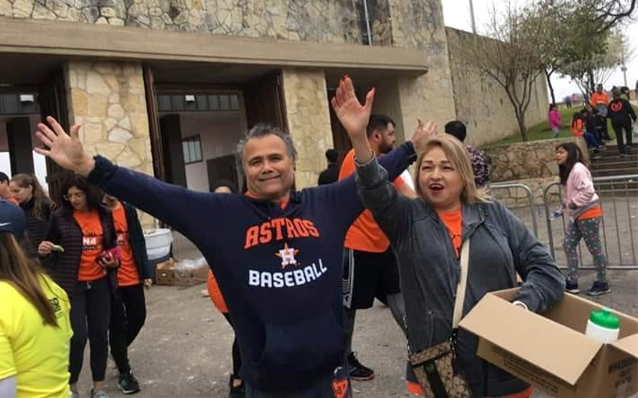 People wearing Astros apparel and orange shirts celebrate with arms raised outside a stone building.