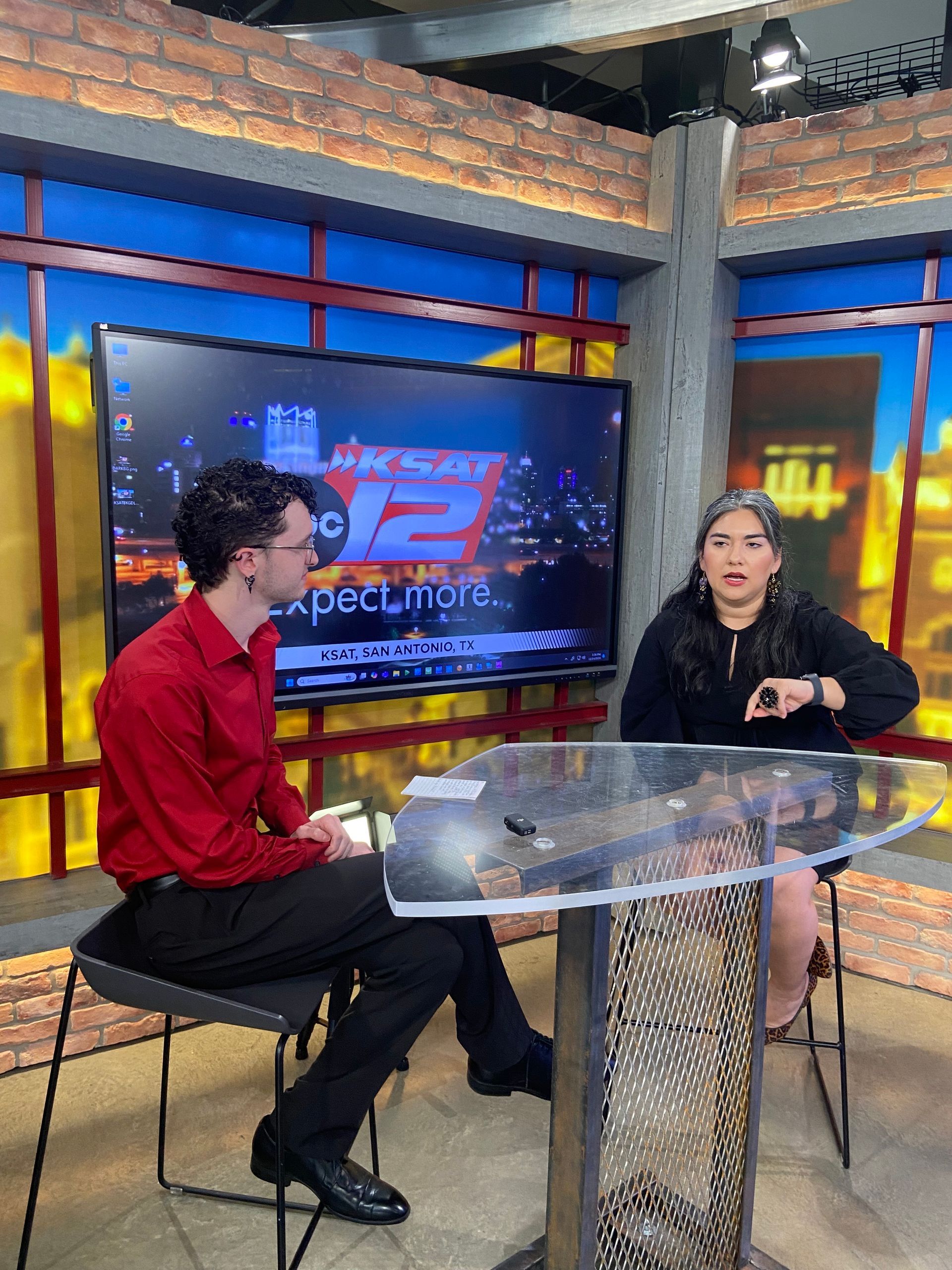 Two people sit at a glass table in a television studio with a large