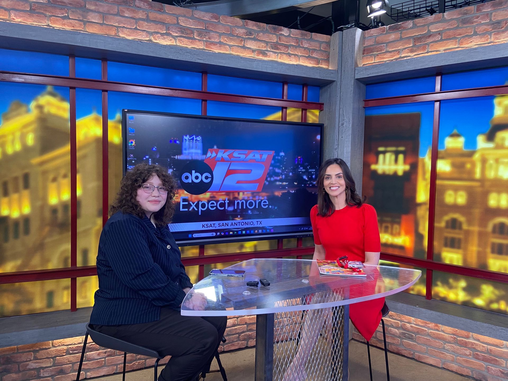 Two people sit at a desk in a television studio in front of an ABC 12 News screen, with city buildings visible behind them.