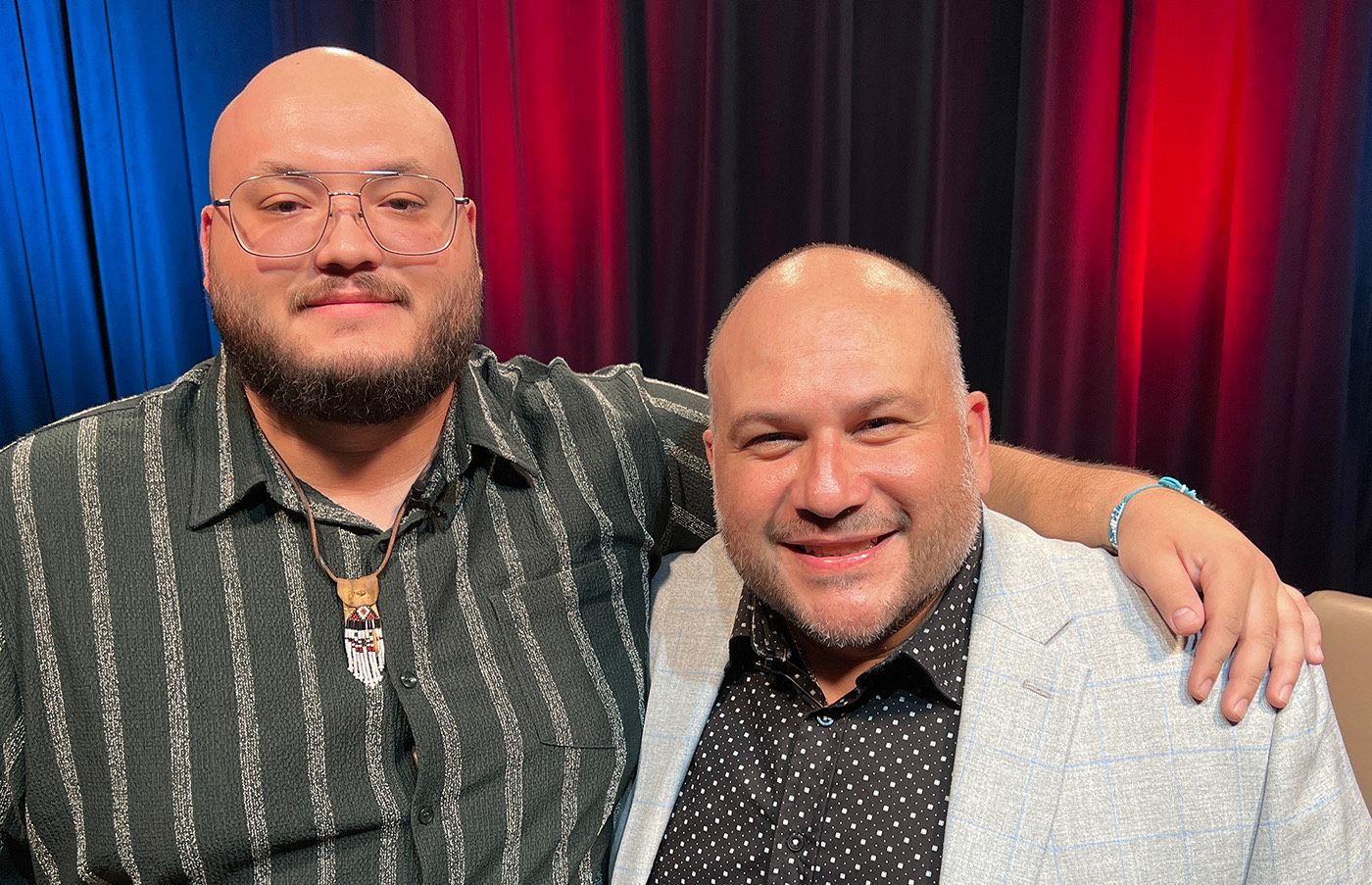 Two bald men smiling with arms around each other in front of a curtain backdrop with blue and red lighting.