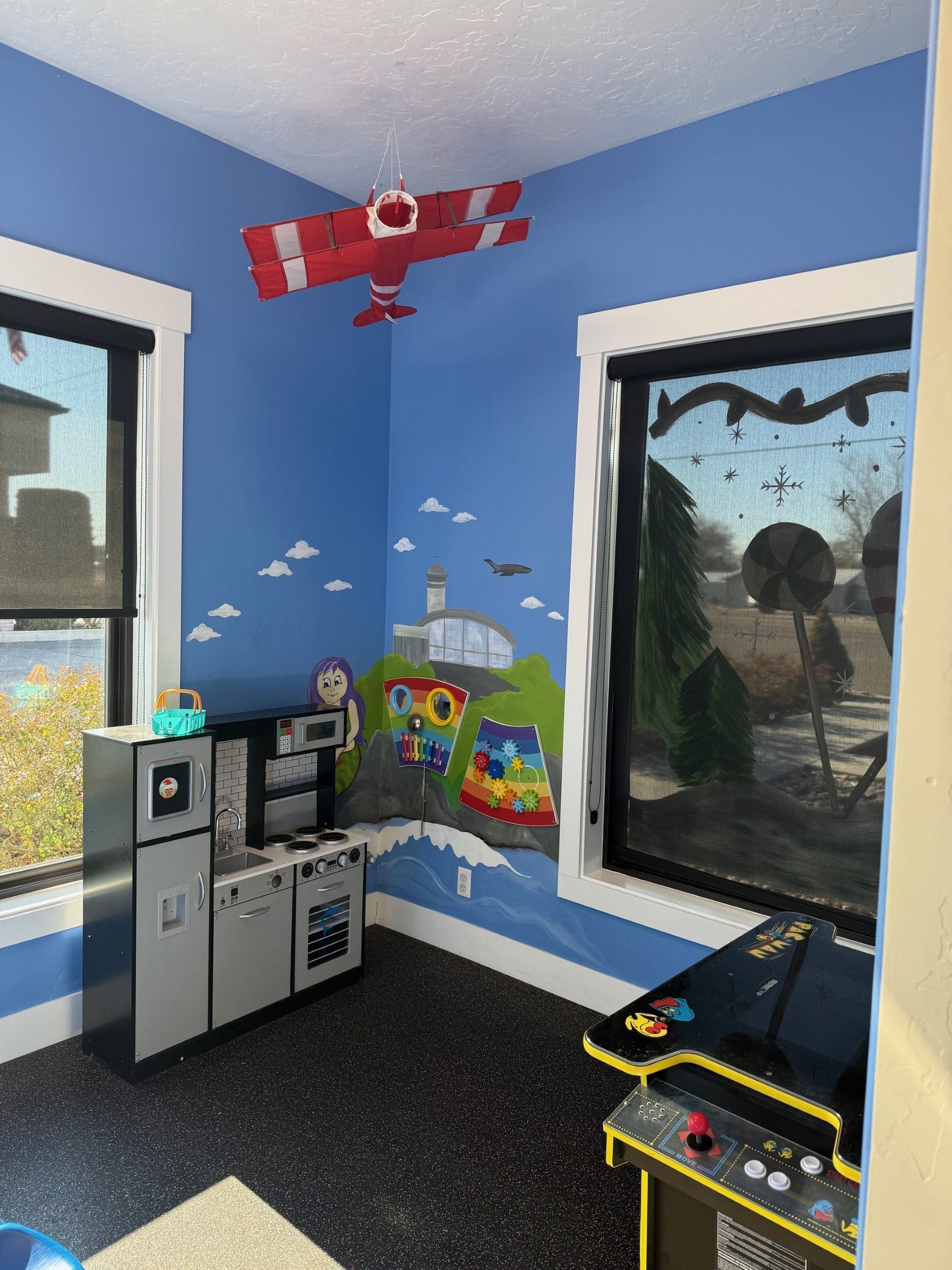 A child's playroom with blue walls, an airplane model hanging from the ceiling, a toy kitchen, and a small arcade game.
