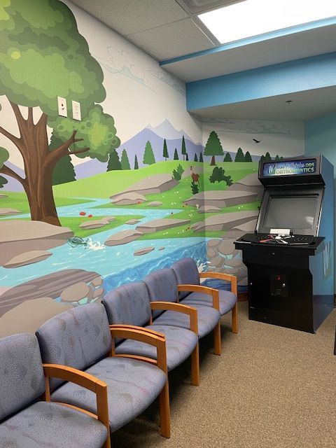 A row of blue chairs in a waiting room with a mural of a tree, stream, and mountains, next to an arcade cabinet.