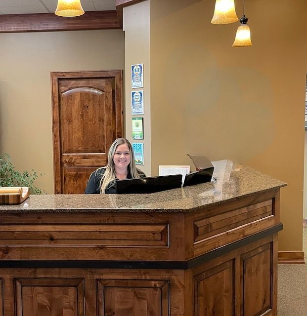 A smiling receptionist sits behind a wood-paneled granite reception desk in a well-lit office with tan walls.