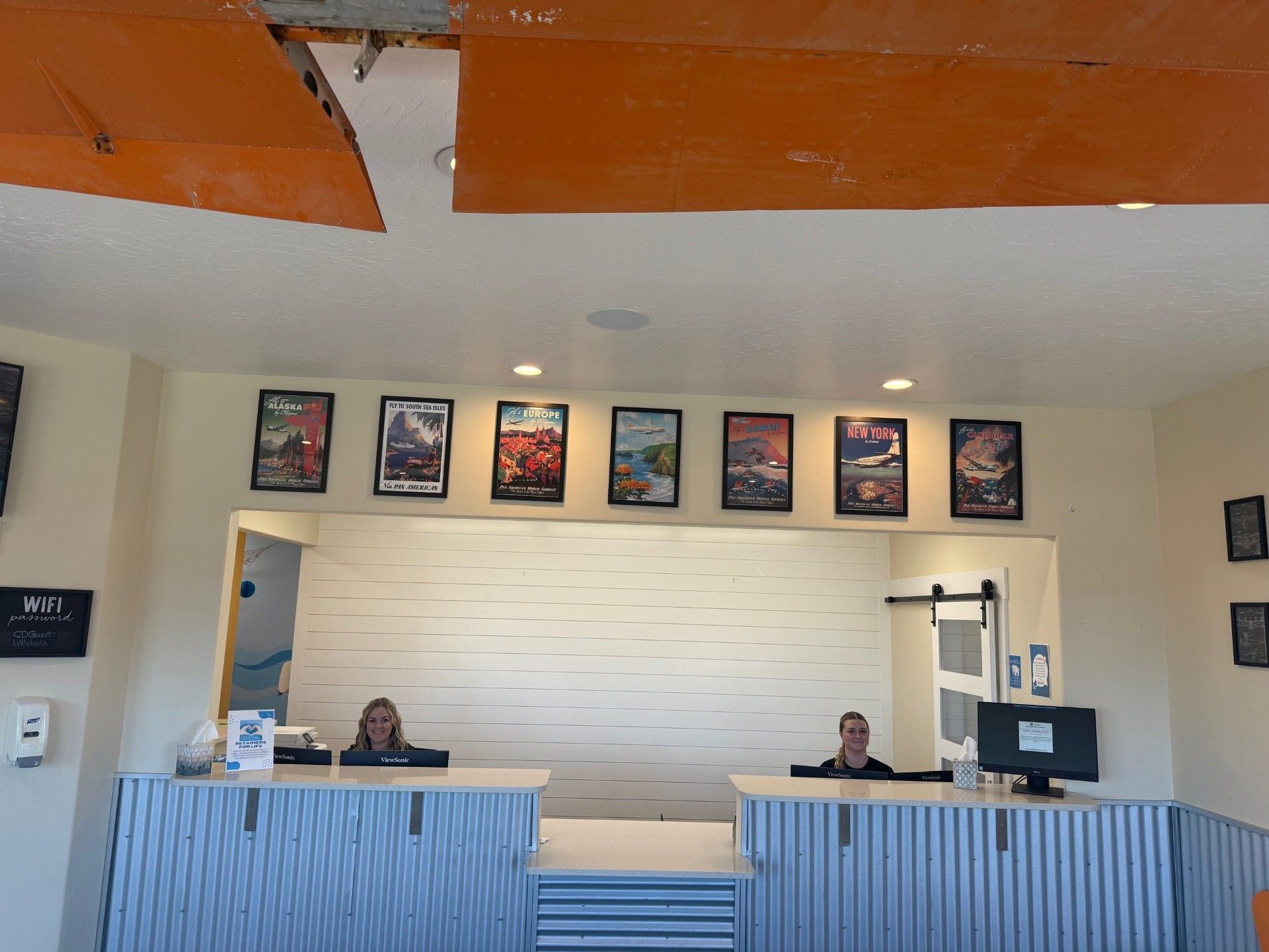 Two employees stand behind a blue-striped reception desk in a brightly lit office with travel-themed wall art.