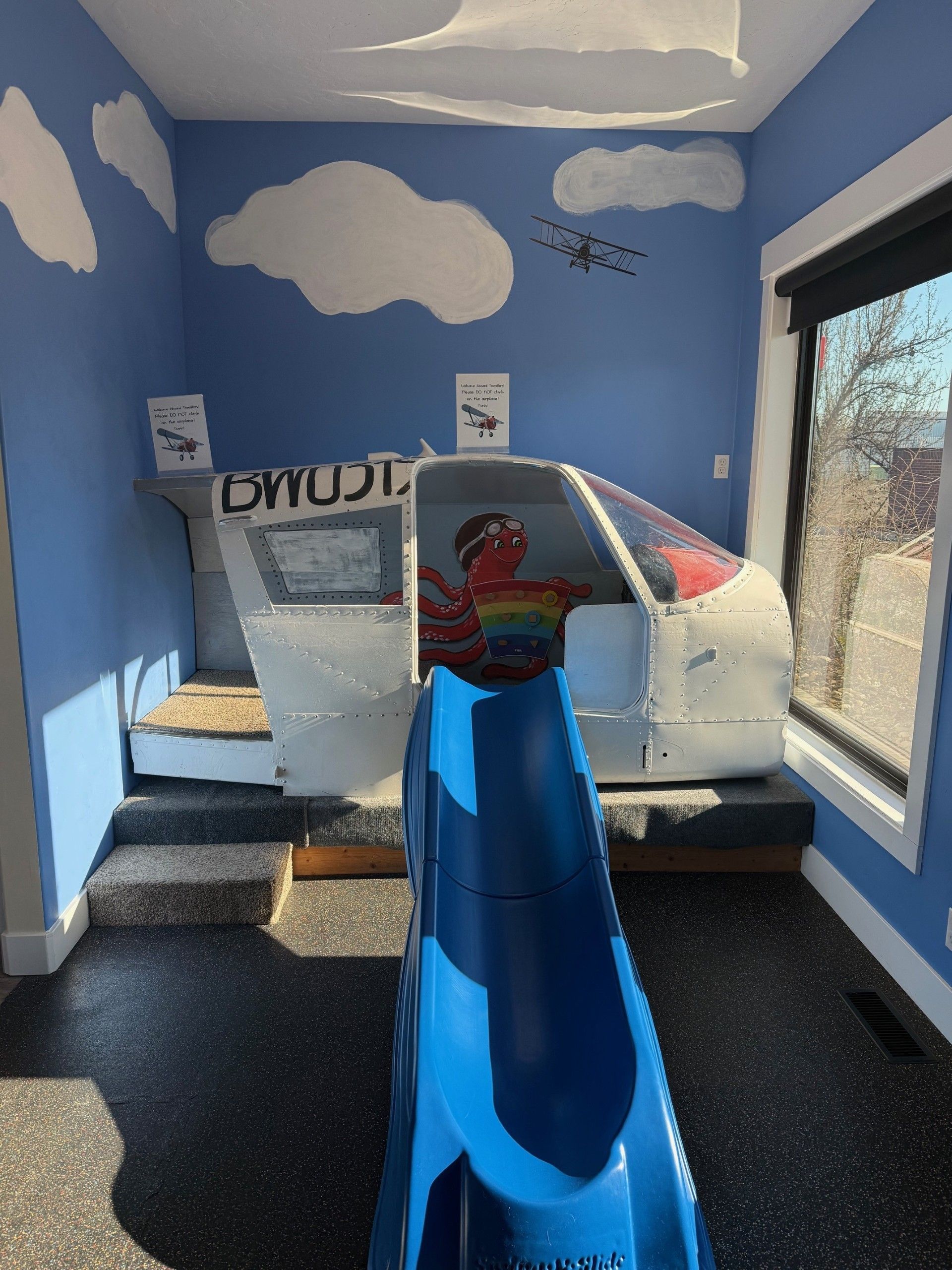 A child's playroom featuring an airplane cockpit built into a wall with a blue slide extending into the room.