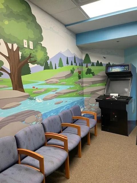 Waiting room with mural of a landscape, blue chairs, and a black arcade game.