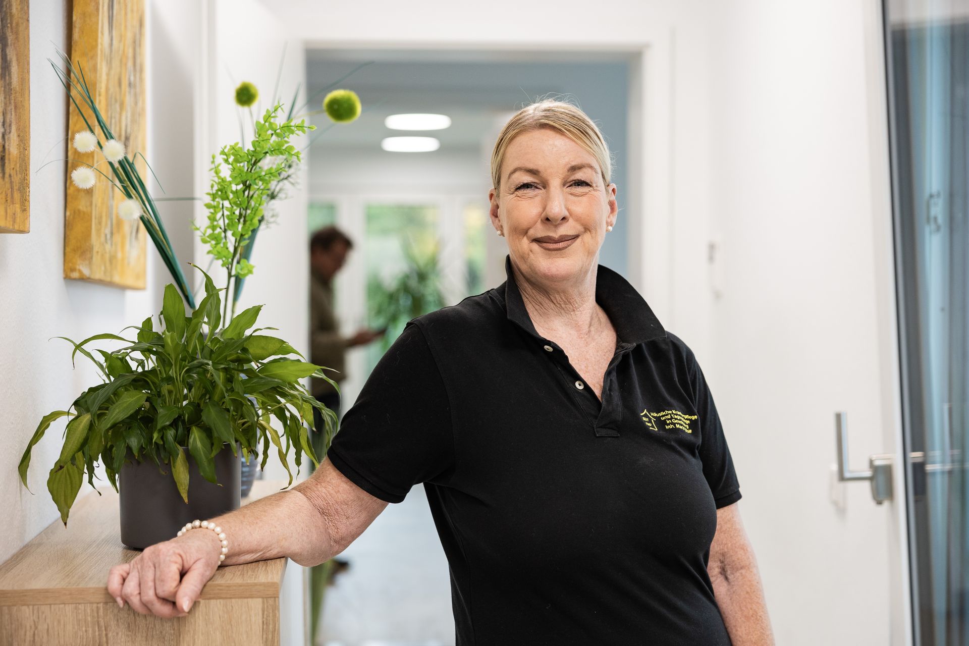 Eine Frau in schwarzem Hemd lächelt in einem Flur und lehnt an einem Holzschrank mit Pflanzen.
