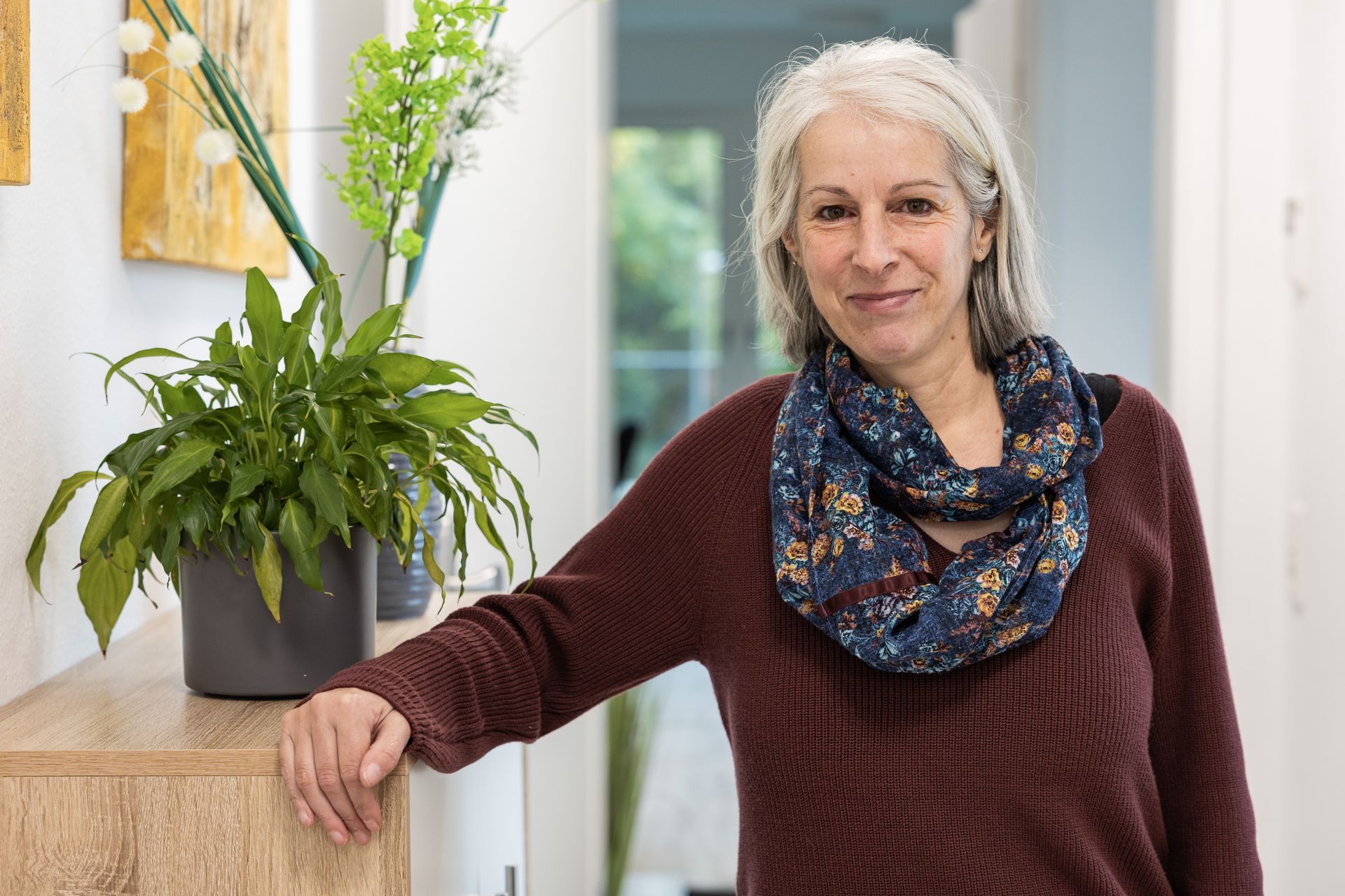 Eine Frau mit grauem Haar, die einen bordeauxroten Pullover lehnt an einer Holztheke neben Pflanzen.