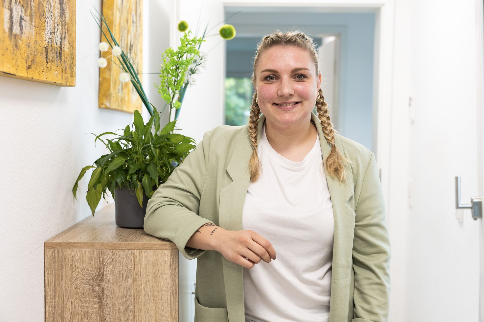 Eine Frau in einem grünen Blazer lächelt neben einem Holzschrank mit einer Pflanze; im Hintergrund ein Flur.