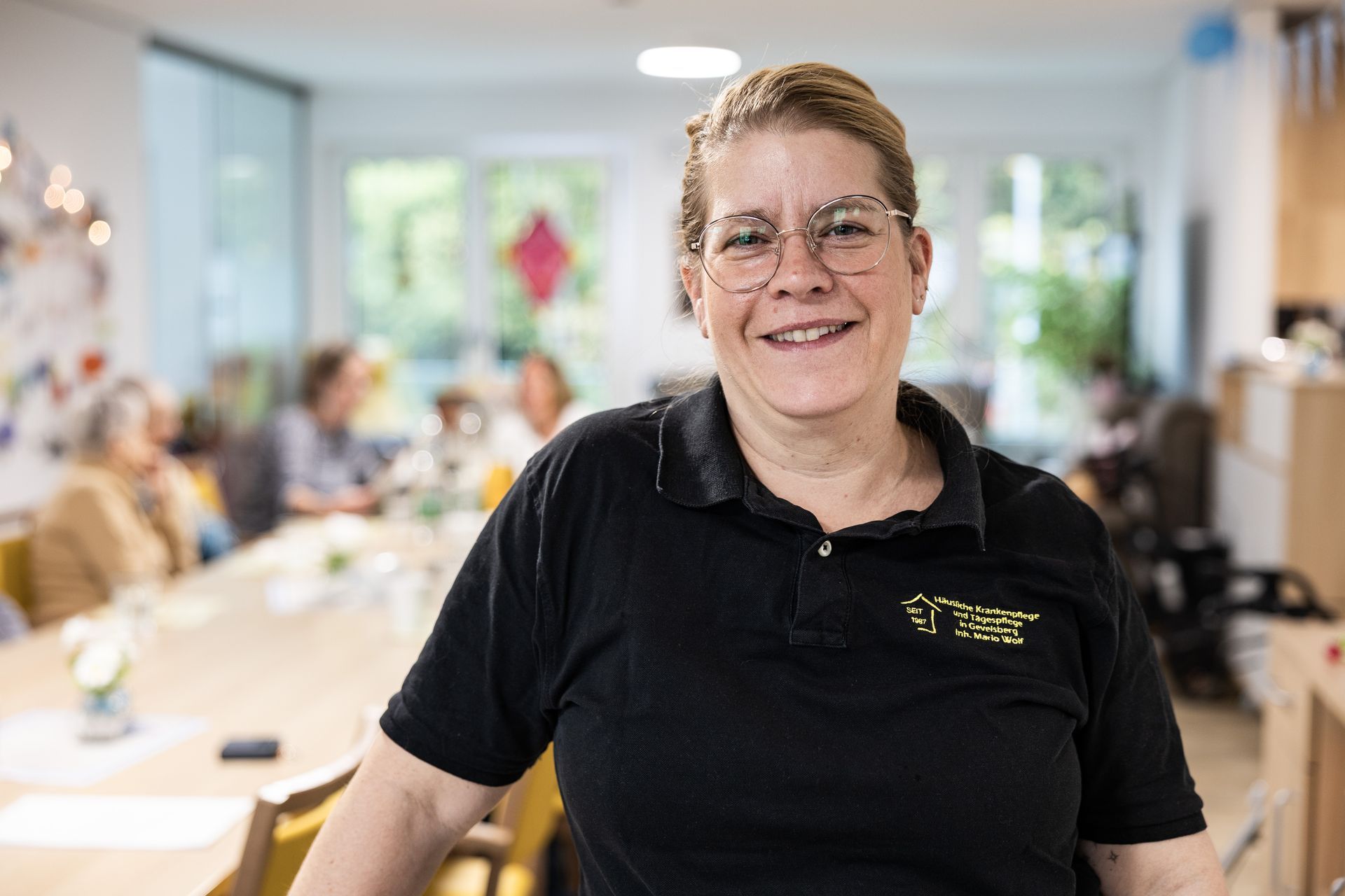 Eine Frau in einem schwarzen Poloshirt lächelt und steht in einem hell erleuchteten Raum.