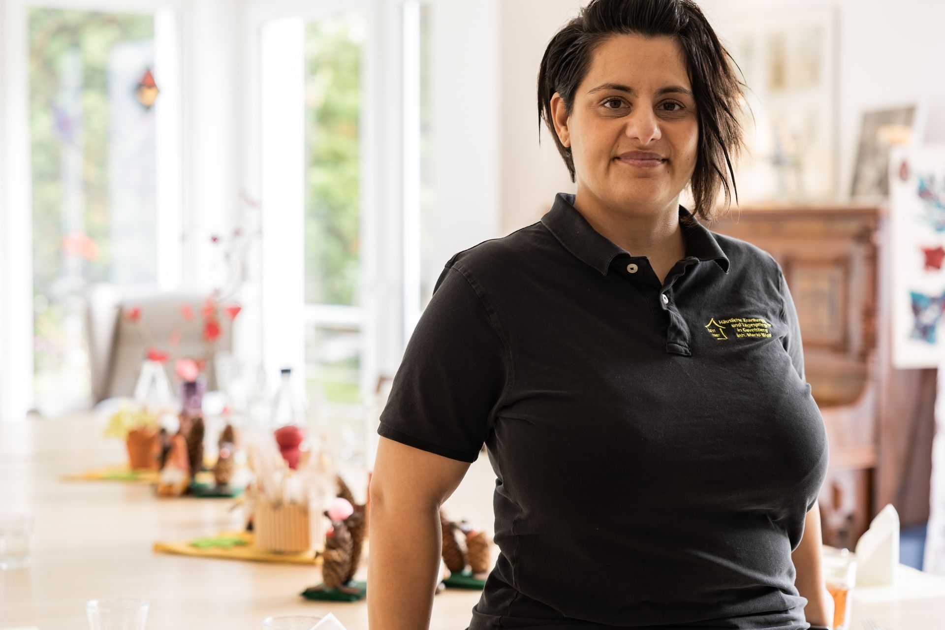Eine Frau in einem schwarzen Poloshirt steht in einem hell erleuchteten Raum neben einem Tisch mit Speisen und Getränken.