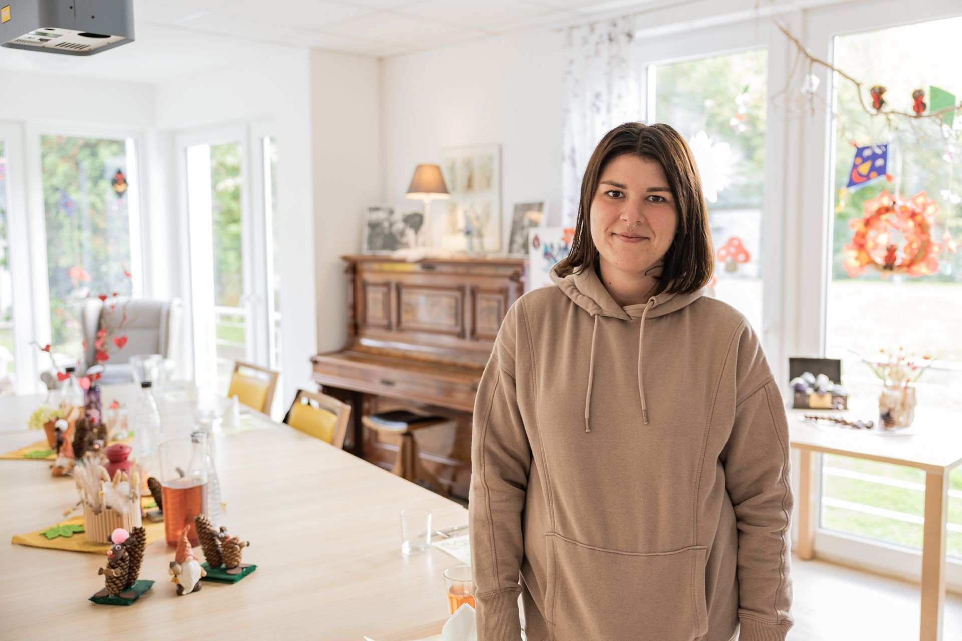 Eine Frau in einem beigen Kapuzenpullover lächelt in einem sonnendurchfluteten Raum.