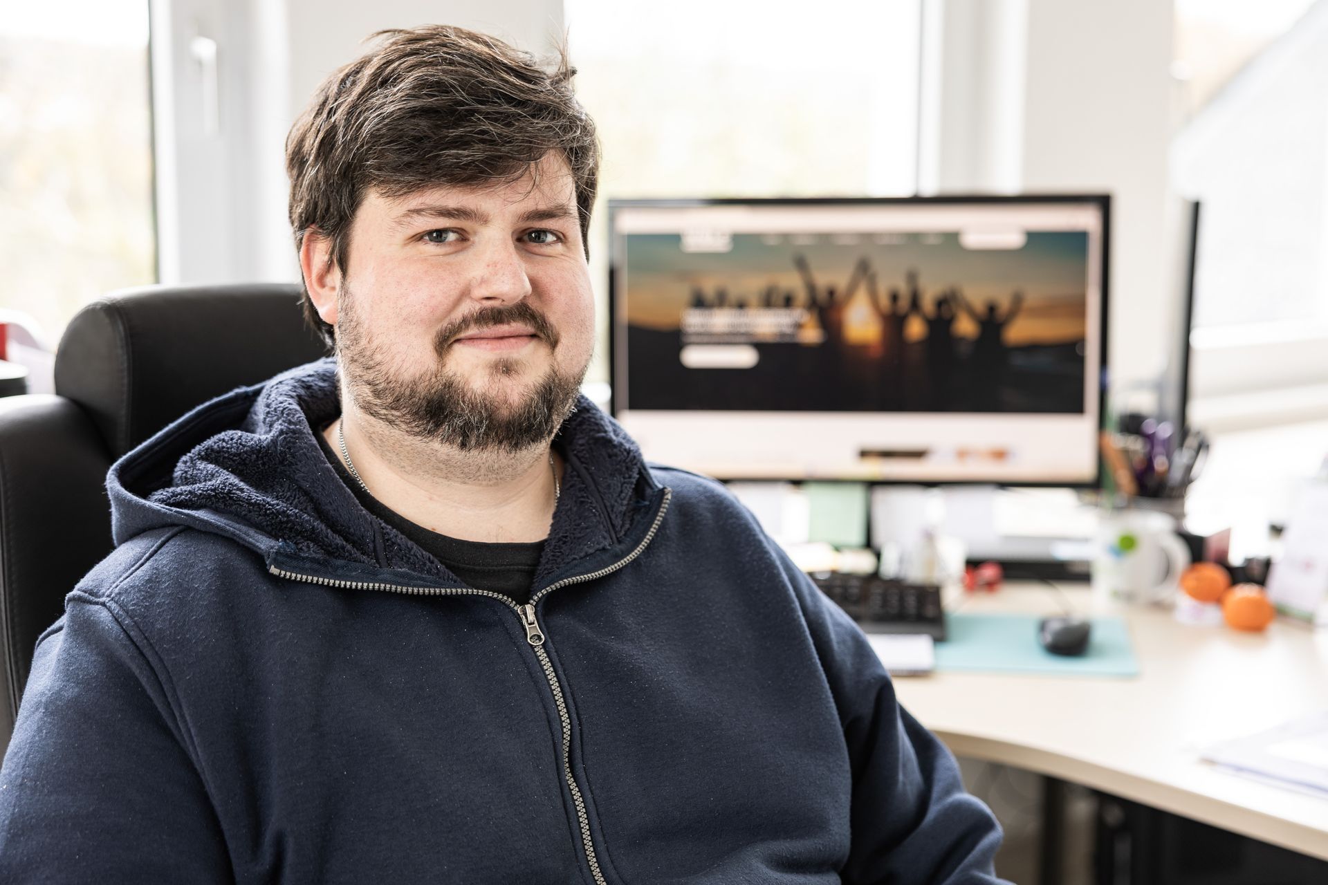 Ein Mann in einem blauen Kapuzenpulli lächelt an einem Schreibtisch, an dem ein Computer eine Webseite anzeigt.