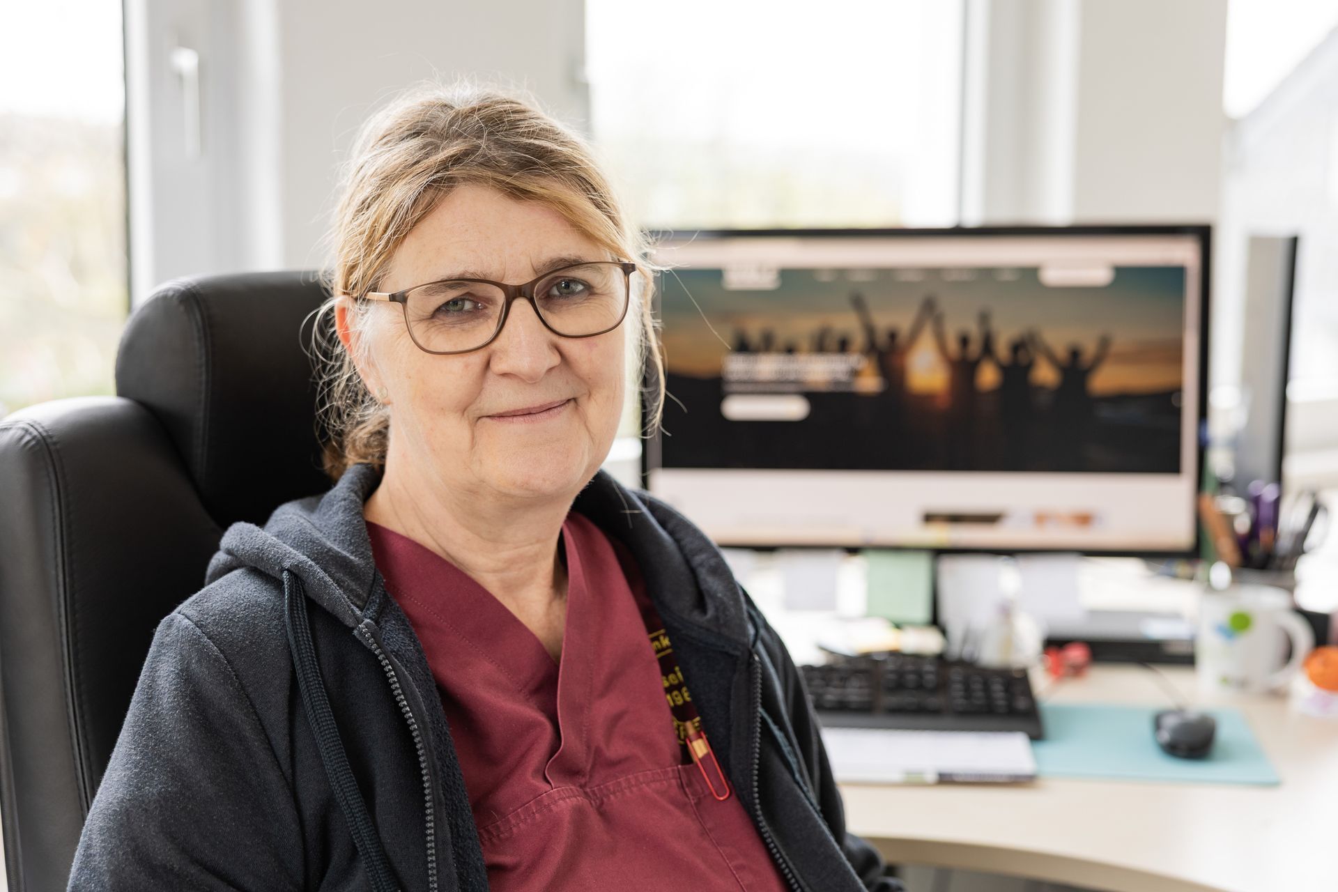 Frau mit Brille sitzt lächelnd an einem Schreibtisch, sie trägt ein rotes Hemd und einen blauen Kapuzenpullover.
