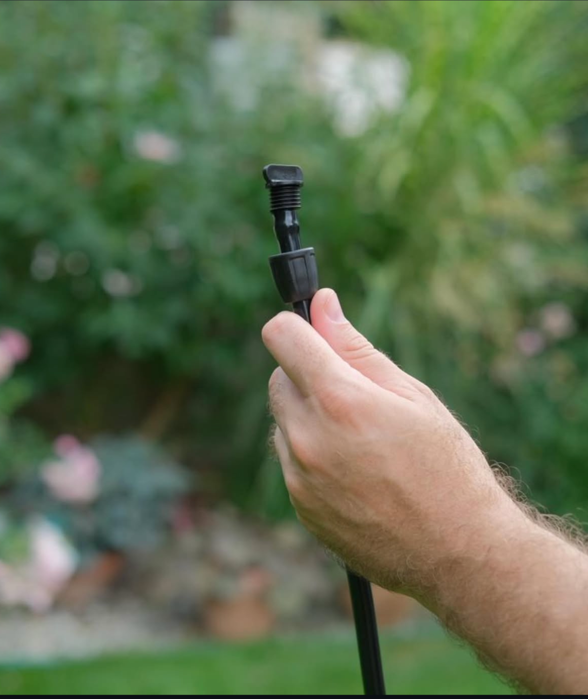 A person holds a small, black irrigation emitter component against a blurred garden background.