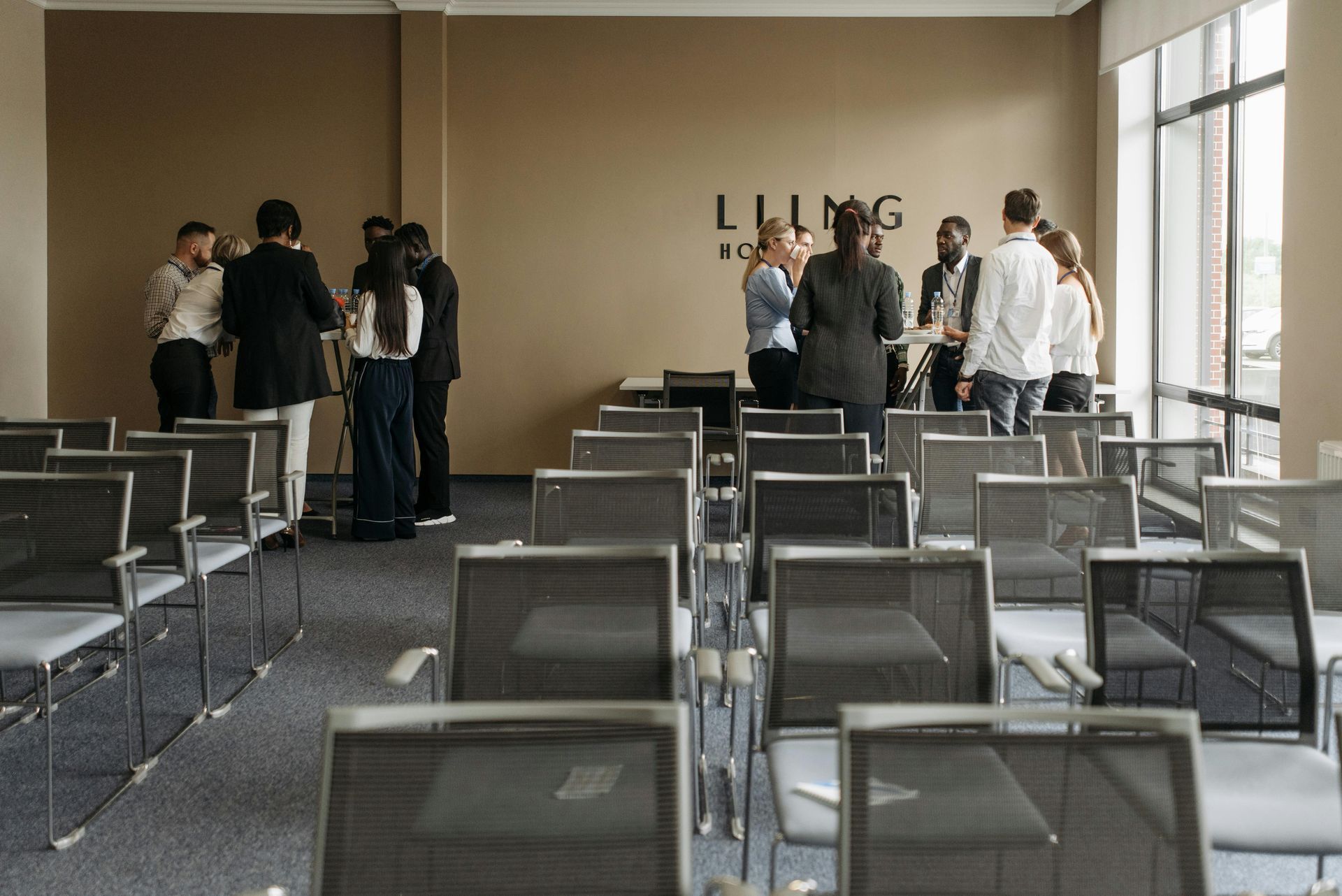Un grupo de personas de pie conversando en un espacio de oficina moderno, con filas de sillas vacías en primer plano.