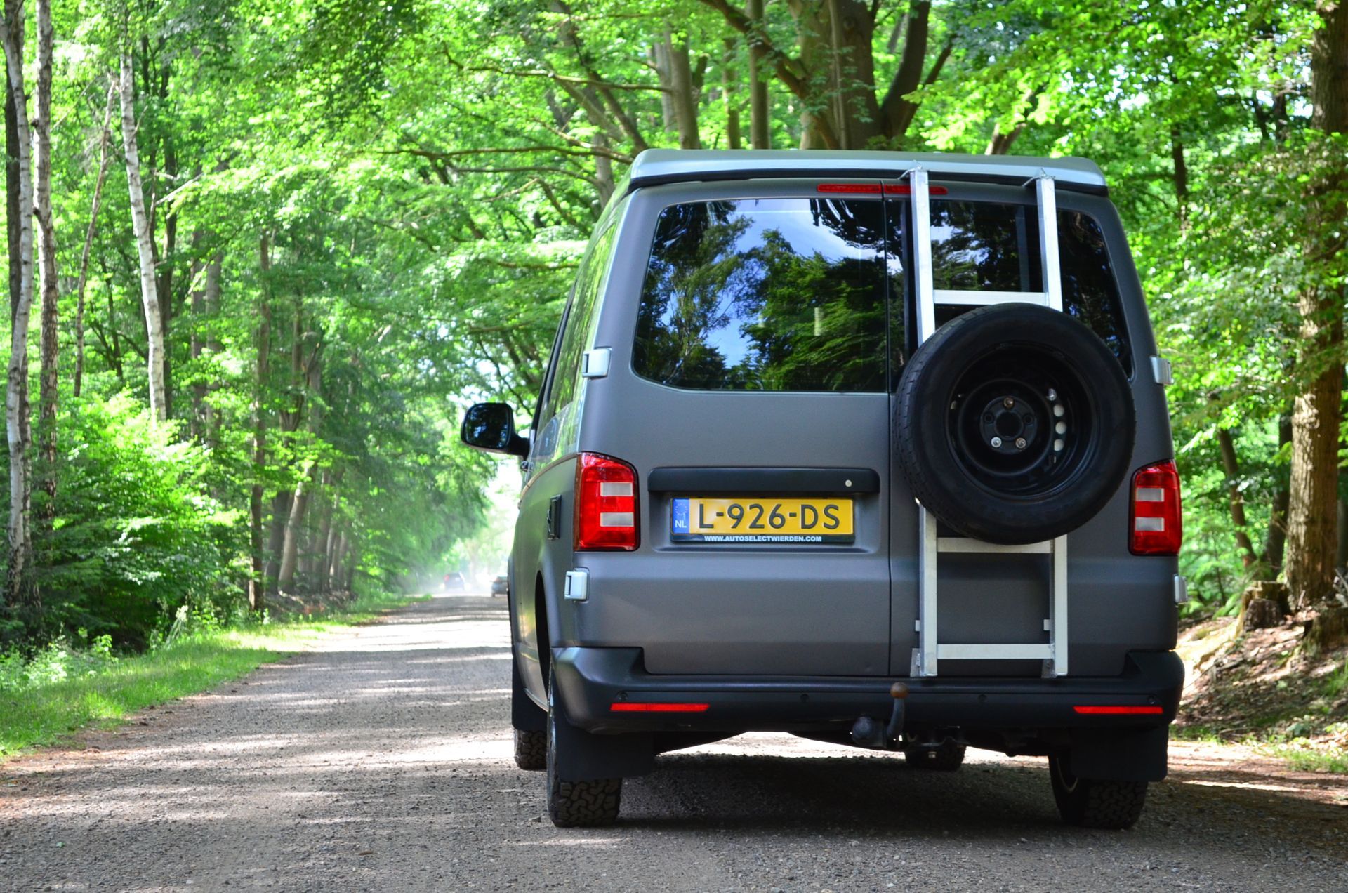 Grijze bestelwagen met reserveband en ladder op een onverharde weg in een bos.