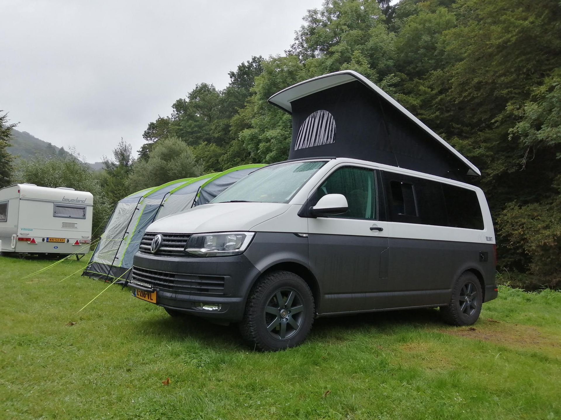 Grijze en witte camperbus met hefdak geparkeerd op het gras naast een tent en een camper.