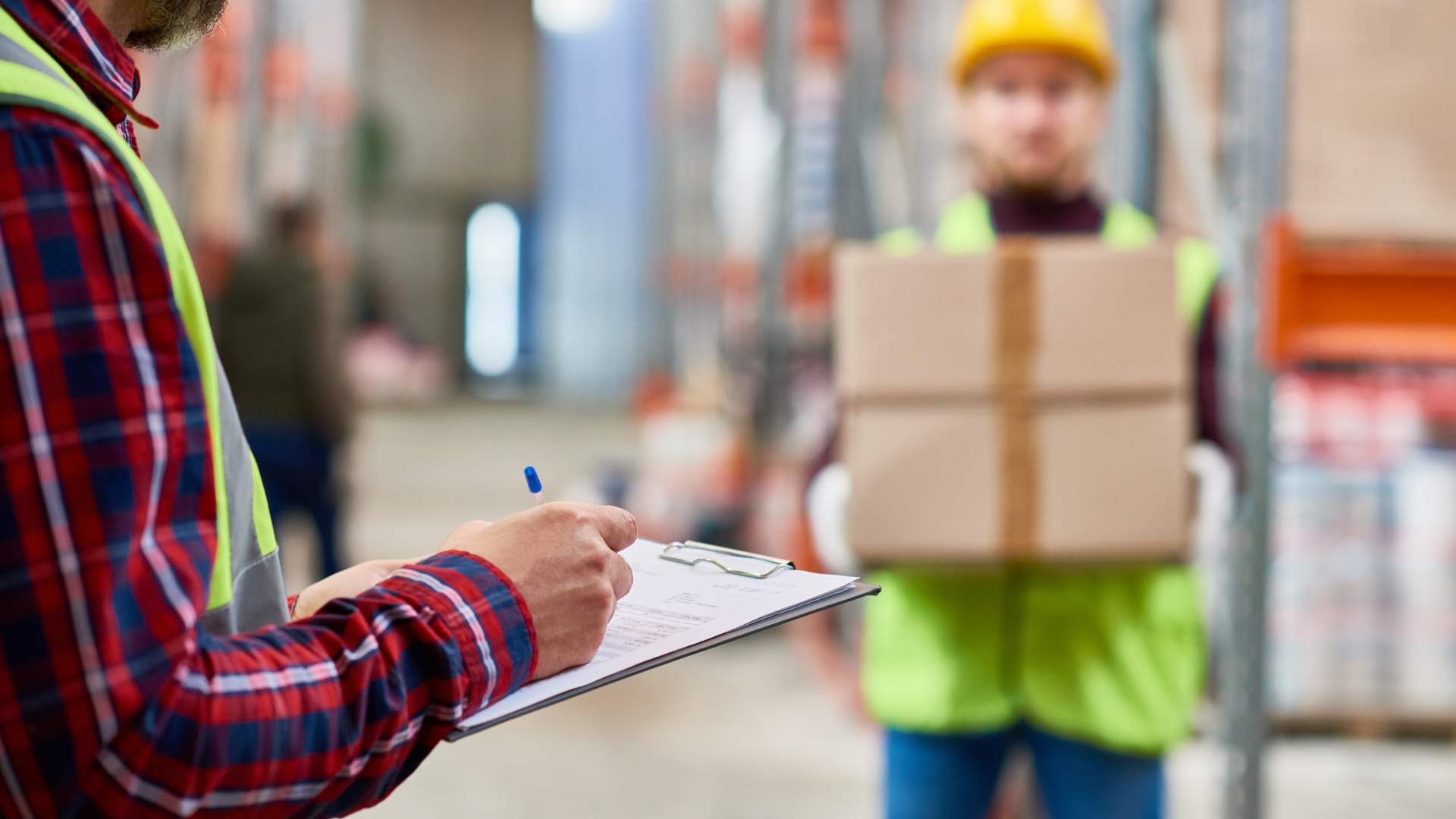 Person taking notes on a clipboard while a worker in a hard hat carries cardboard boxes in a warehouse aisle