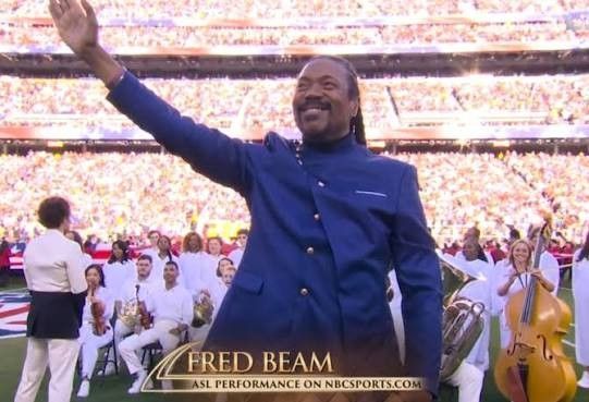 Fred Beam waving on a football field during a stadium ceremony, wearing a blue suit, with a cheering crowd behind him