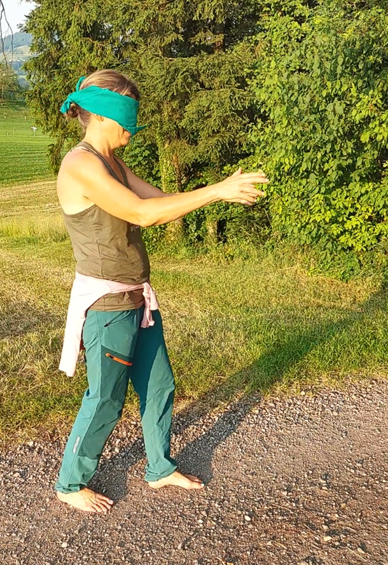 Person mit verbundenen Augen im Freien, Arme ausgestreckt, bekleidet mit grüner Hose, Oberteil und Bandana.