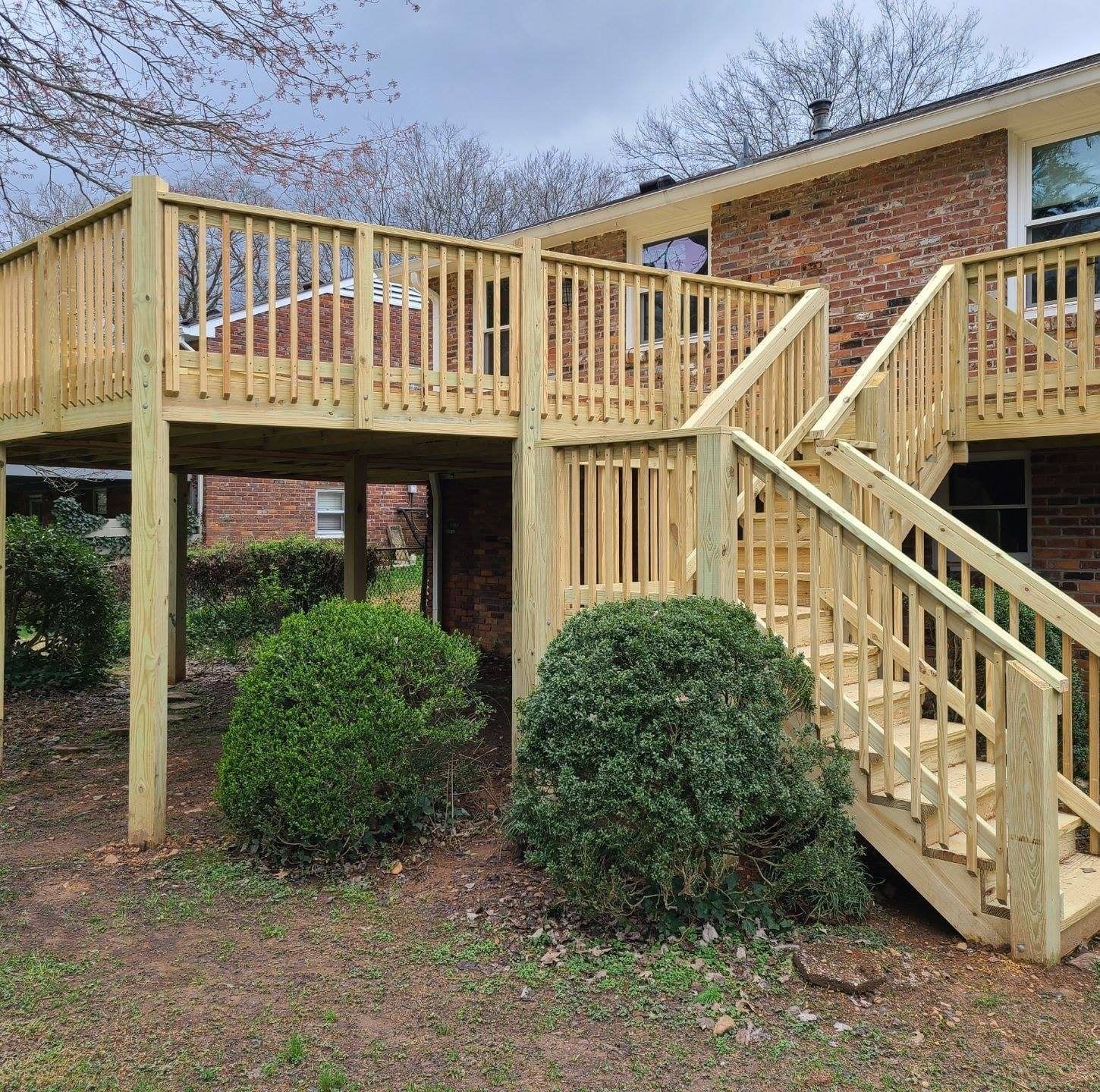 Wooden deck and stairs with railings beside a brick house above trimmed shrubs