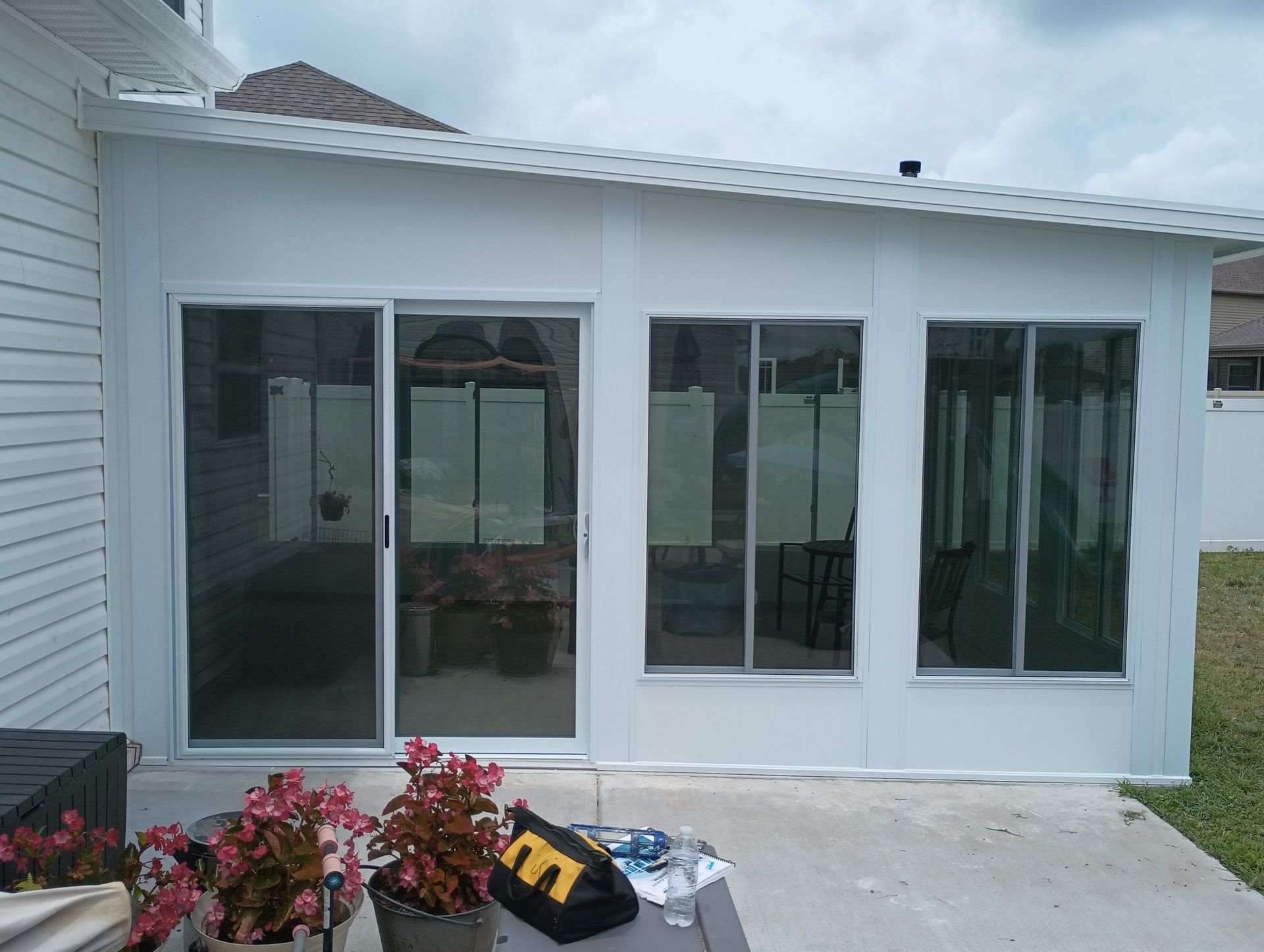 White sunroom with large glass sliding doors, white siding, and potted flowers on the patio.