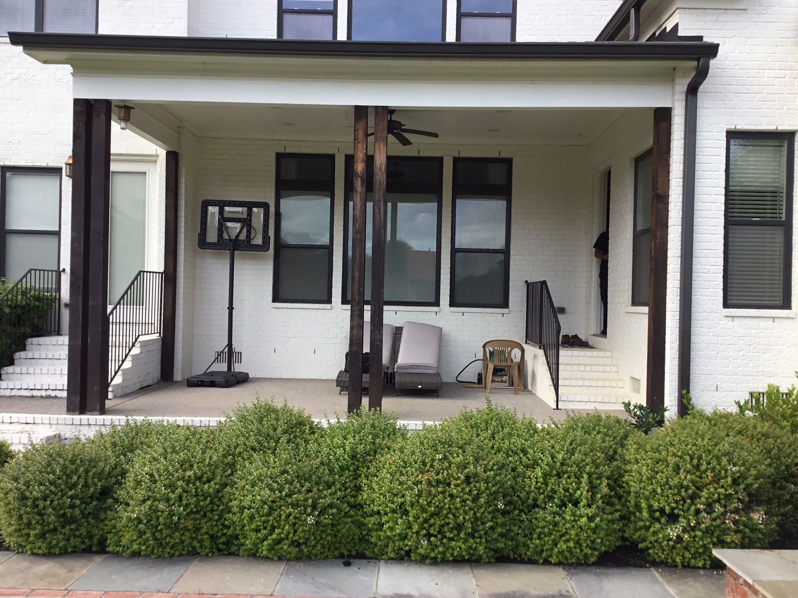 Front porch of a white house with black trim, chairs, and a hedge in front