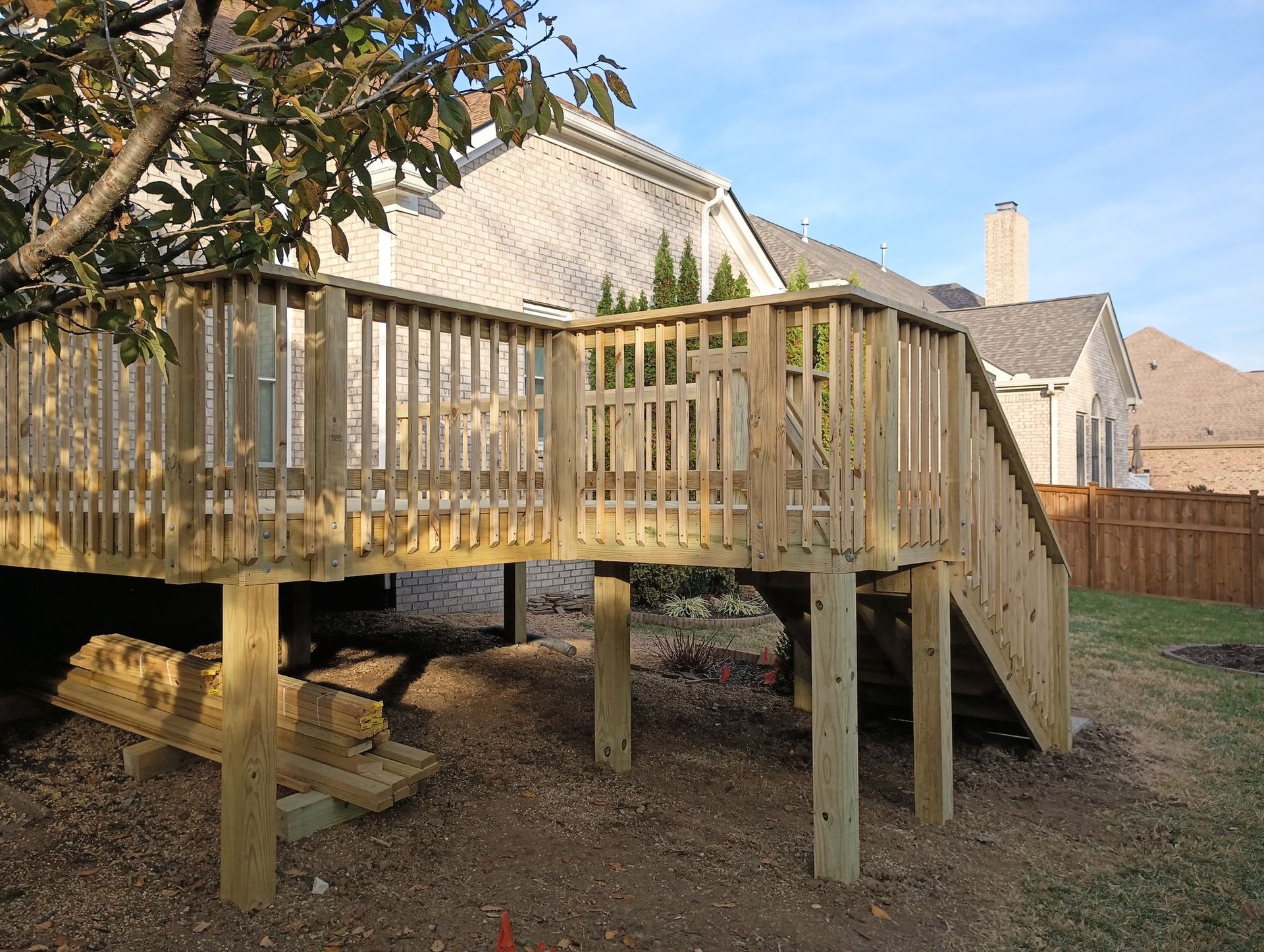 Wooden raised deck with railings and stairs in a backyard, beside a house and fence.
