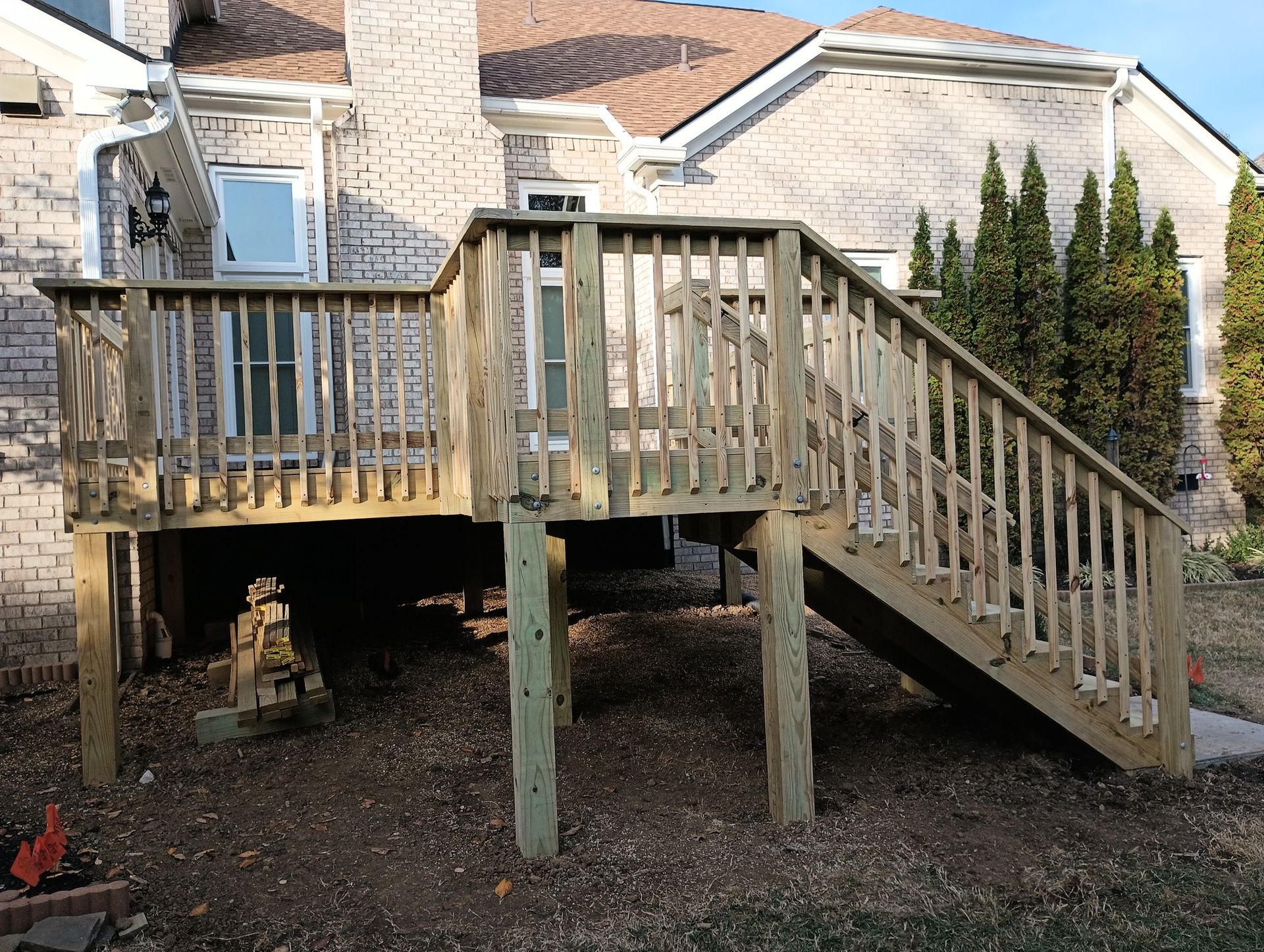 Wooden deck and stairs on stilts beside a stone house, with a gravel yard below.