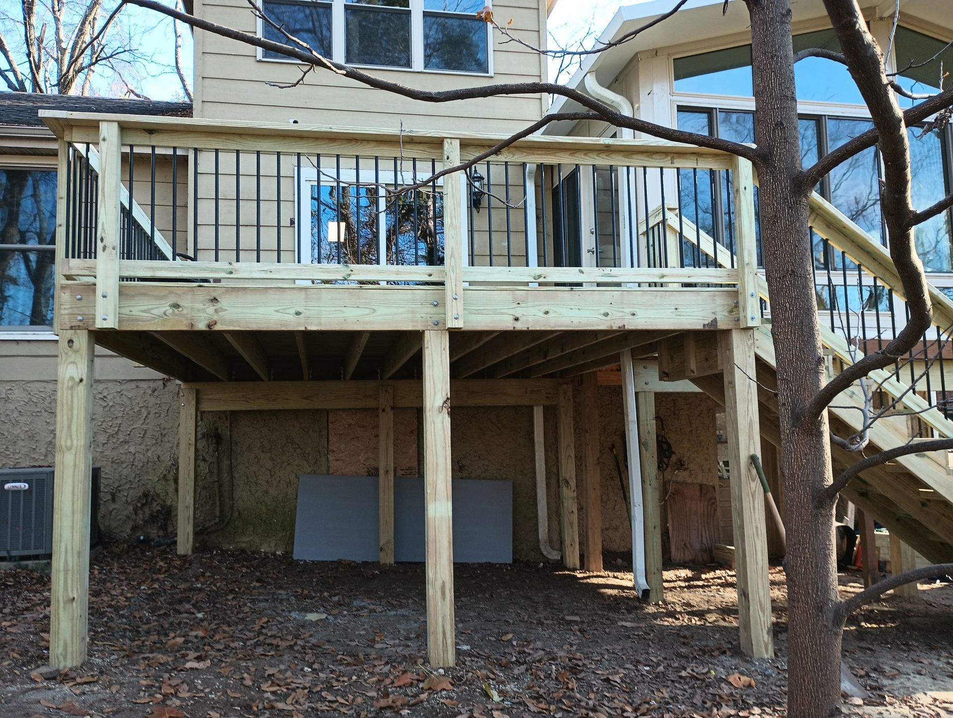 Backyard deck supported by wooden posts beneath a house, with leafless trees and a brick wall nearby