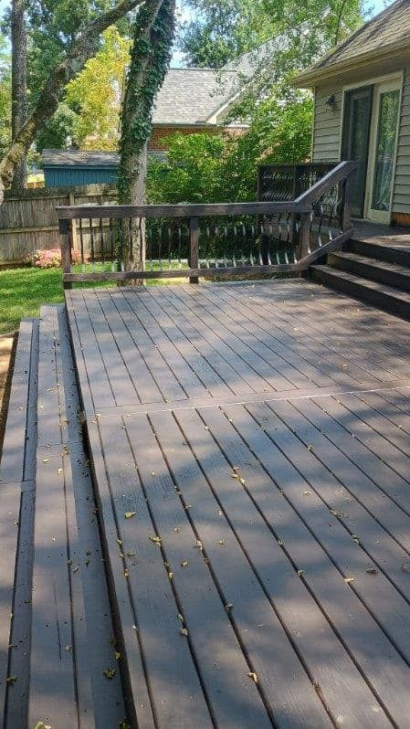 Wooden deck with railing beside a house, shaded by trees and scattered with leaves.