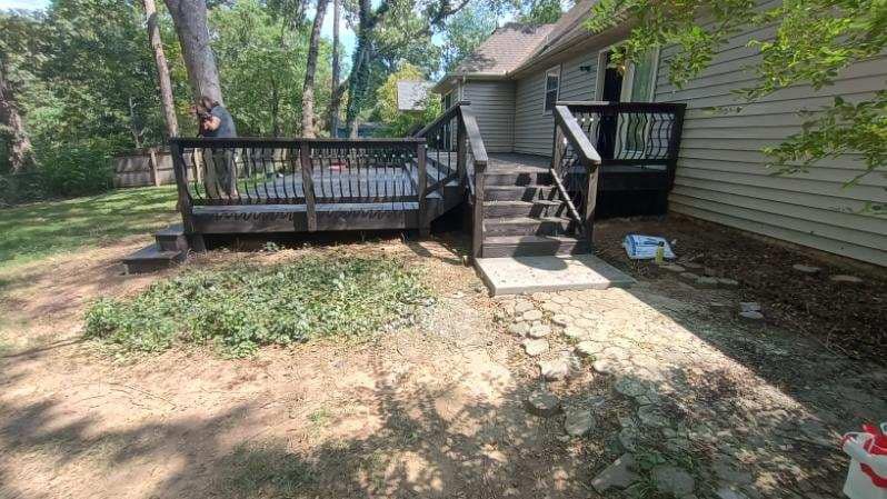 Backyard deck with stairs and a paved path beside a house, surrounded by trees and dirt ground
