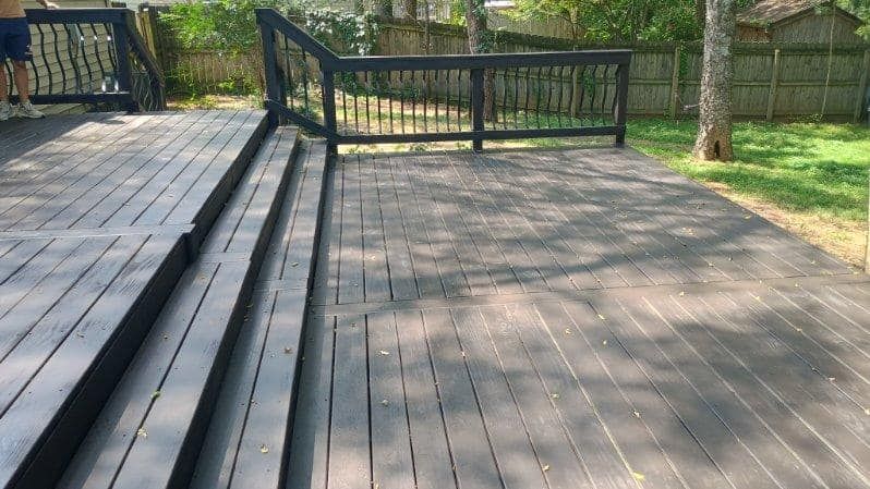 Wooden deck walkway with black railings beside a fenced yard and grassy area.