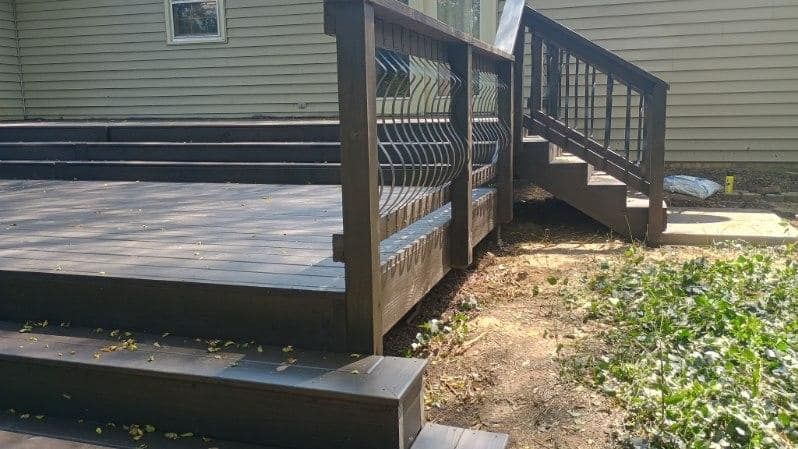 Wooden deck and short stairs beside a beige house, with black railing and a patchy dirt path below.