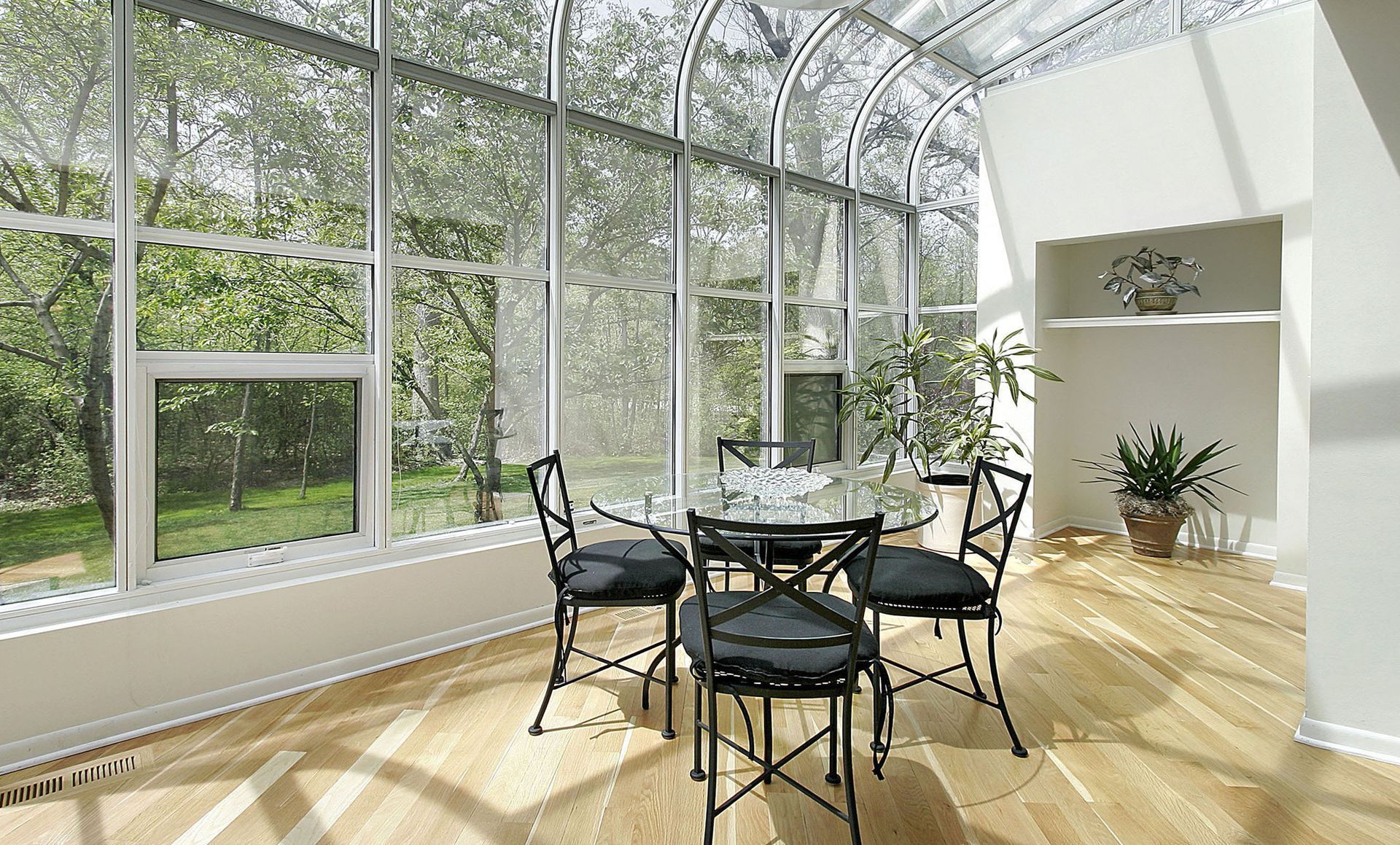 Sunlit glass conservatory with black chairs and round table on a wooden floor, overlooking greenery