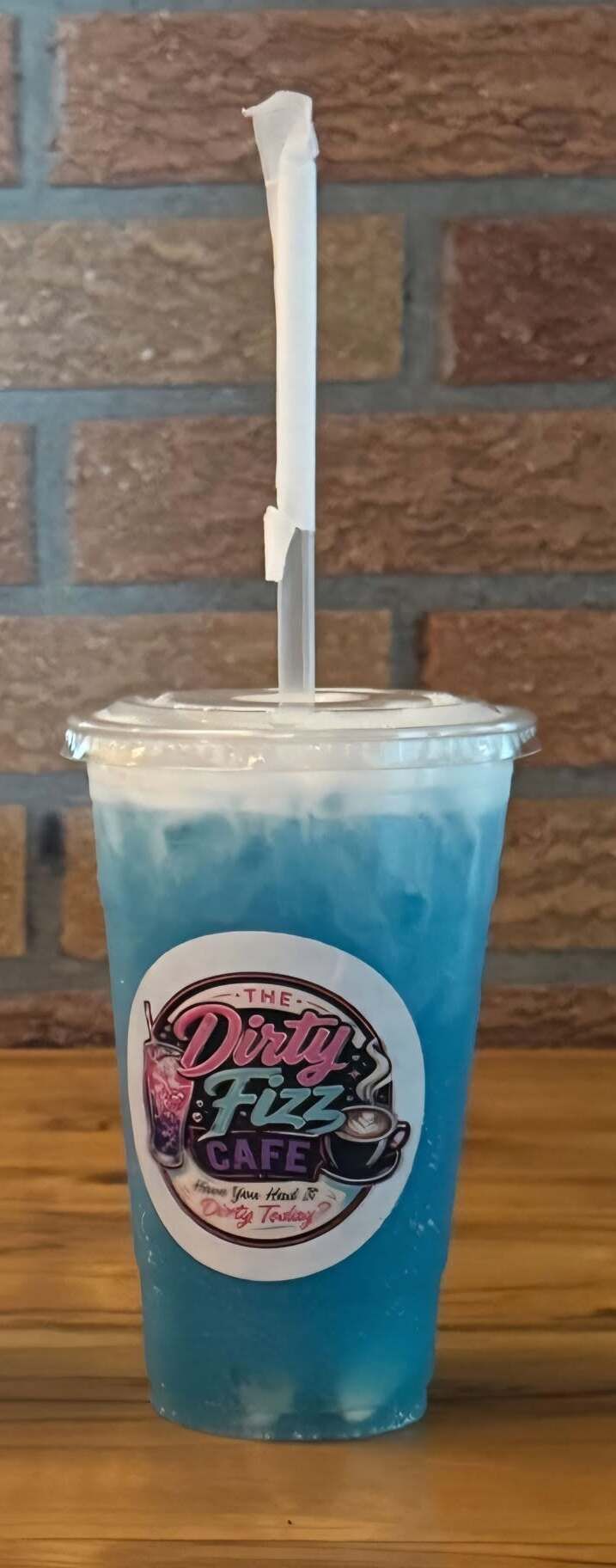 Blue iced drink in a clear plastic cup with a straw, labeled Dirty Fizz Cafe, against a brick wall